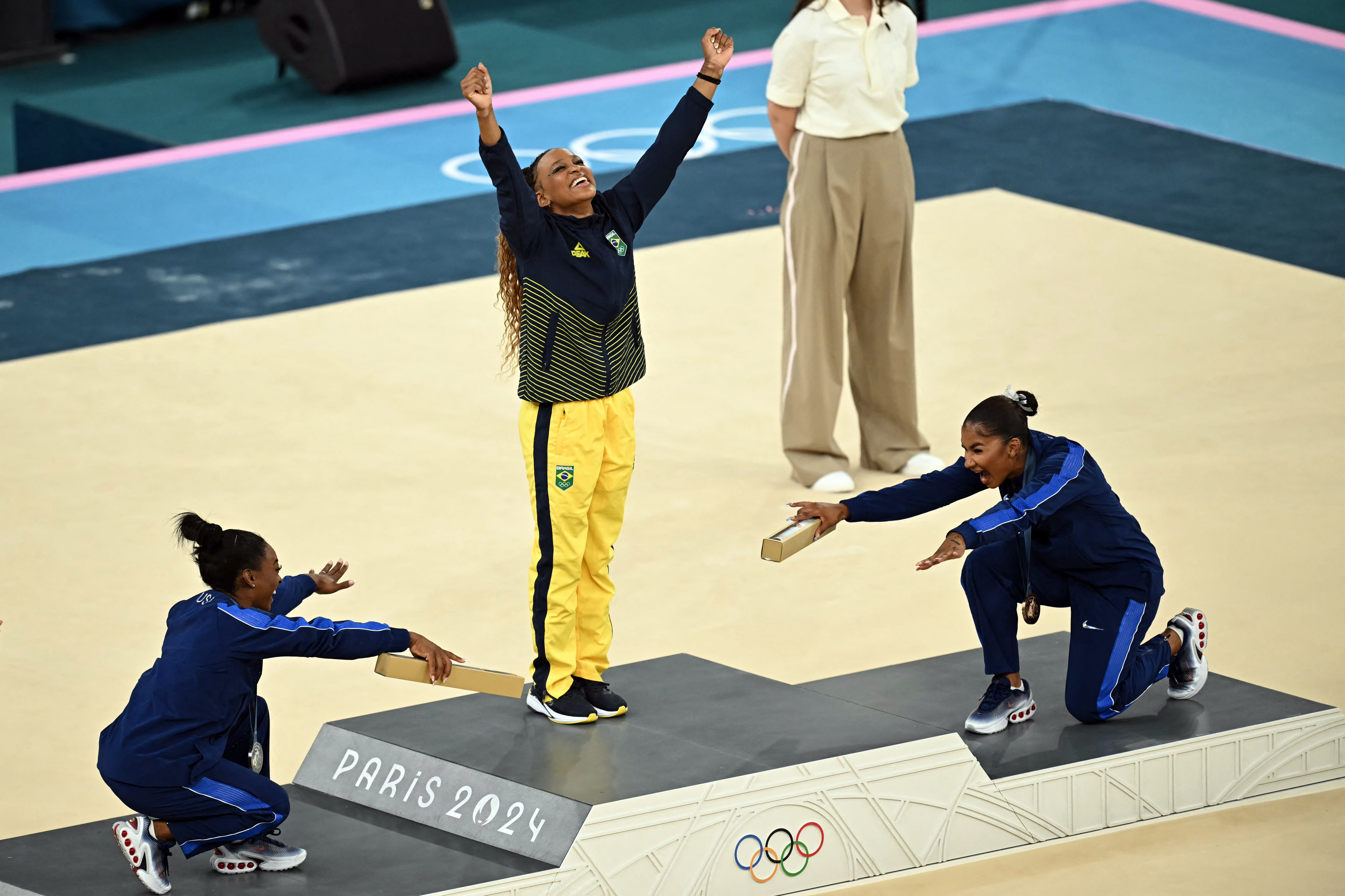 Athletes celebrate on a podium at the 2024 Paris Olympics gymnastics event. One athlete stands with arms raised, while two others kneel on either side, pointing to her
