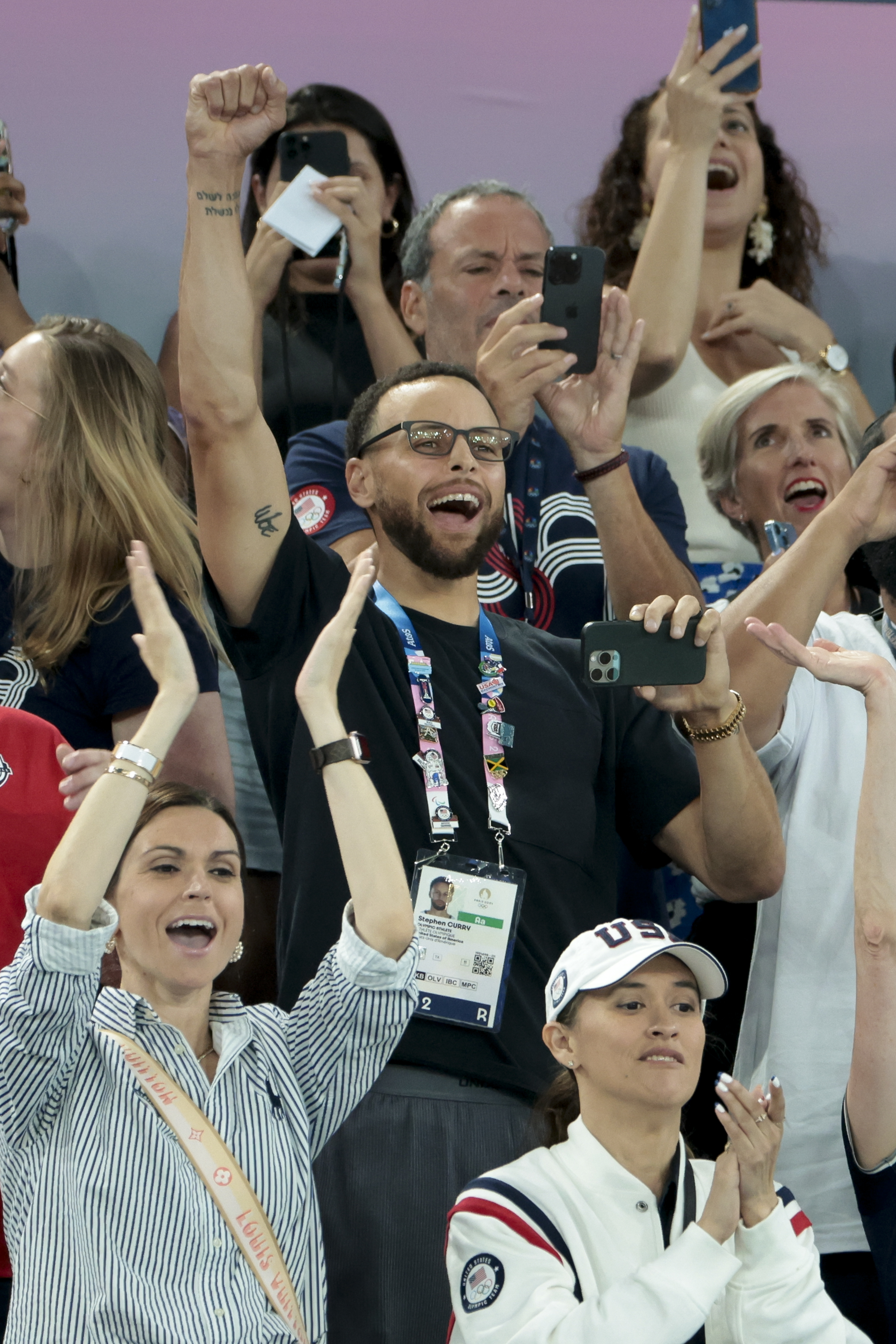 Stephen Curry cheering with fans at an event, wearing a credential lanyard and holding a phone. Excited onlookers are taking photos and celebrating around him