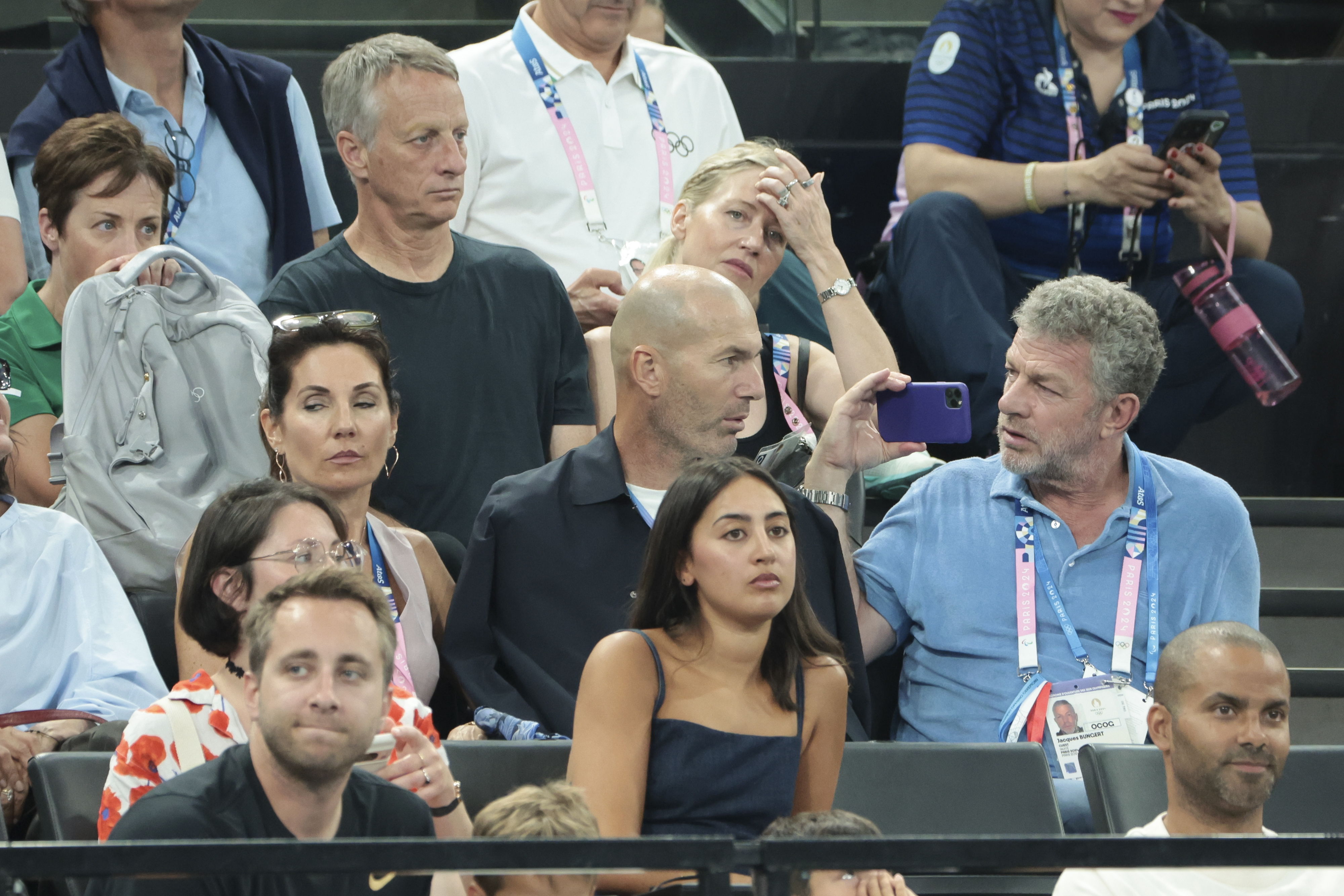 Zinedine Zidane at an event, surrounded by attendees. A man to his left is holding a phone, seemingly taking a photo