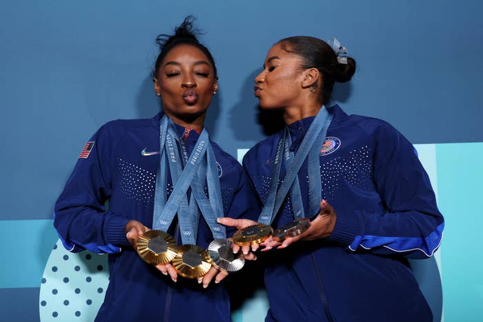Simone Biles and Jordan Chiles celebrate holding multiple Olympic medals while posing playfully in navy athletic jackets