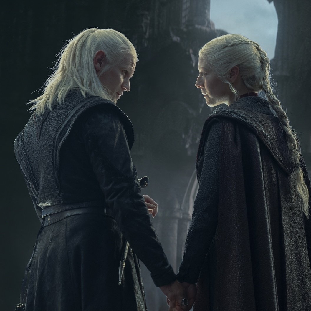 Dos personajes de cabello blanco y trenzado, Geralt de Rivia y Rhaenyra Targaryen, se miran fijamente en un ambiente oscuro y medieval, sosteniendo espadas