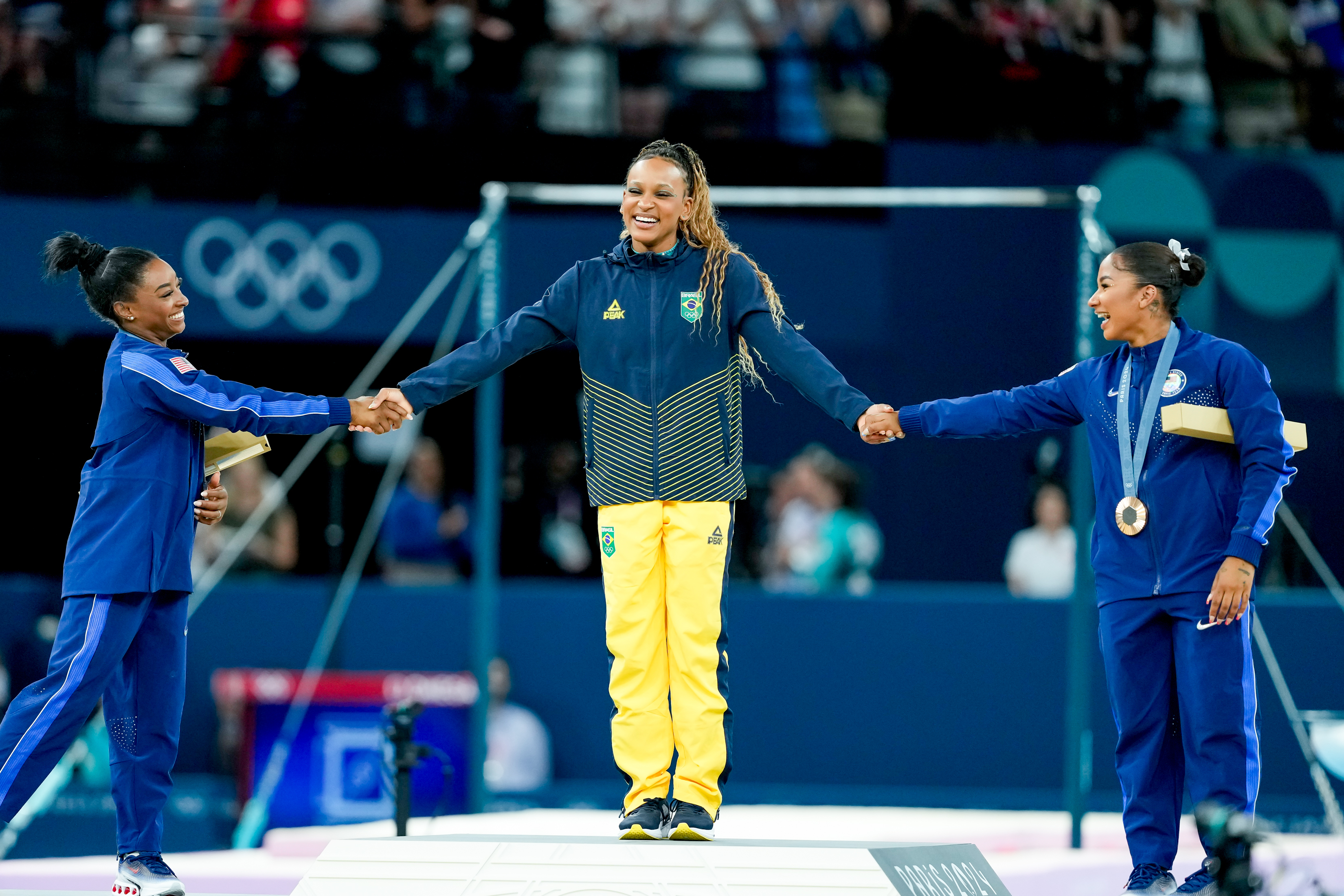 Olympic medalists on the podium: Rebeca Andrade (center) in tracksuit, Flavia Saraiva (left) shakes hands, and Daniele Hypolito (right) with her medal also shakes hands