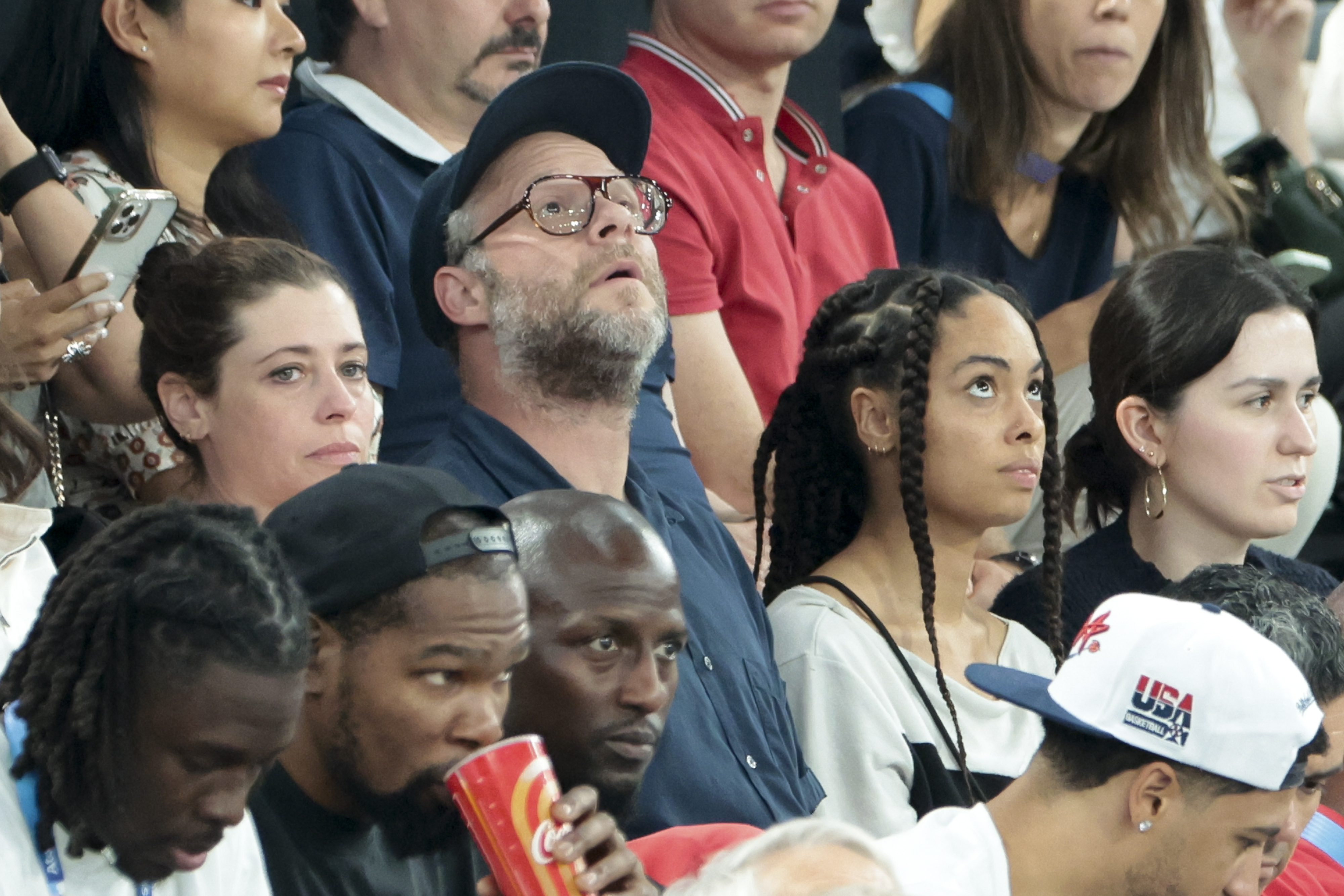Seth Rogen in a crowd at a sports event, wearing glasses and a baseball cap. He's surrounded by spectators, including a woman with braided hair and a man drinking from a large cup
