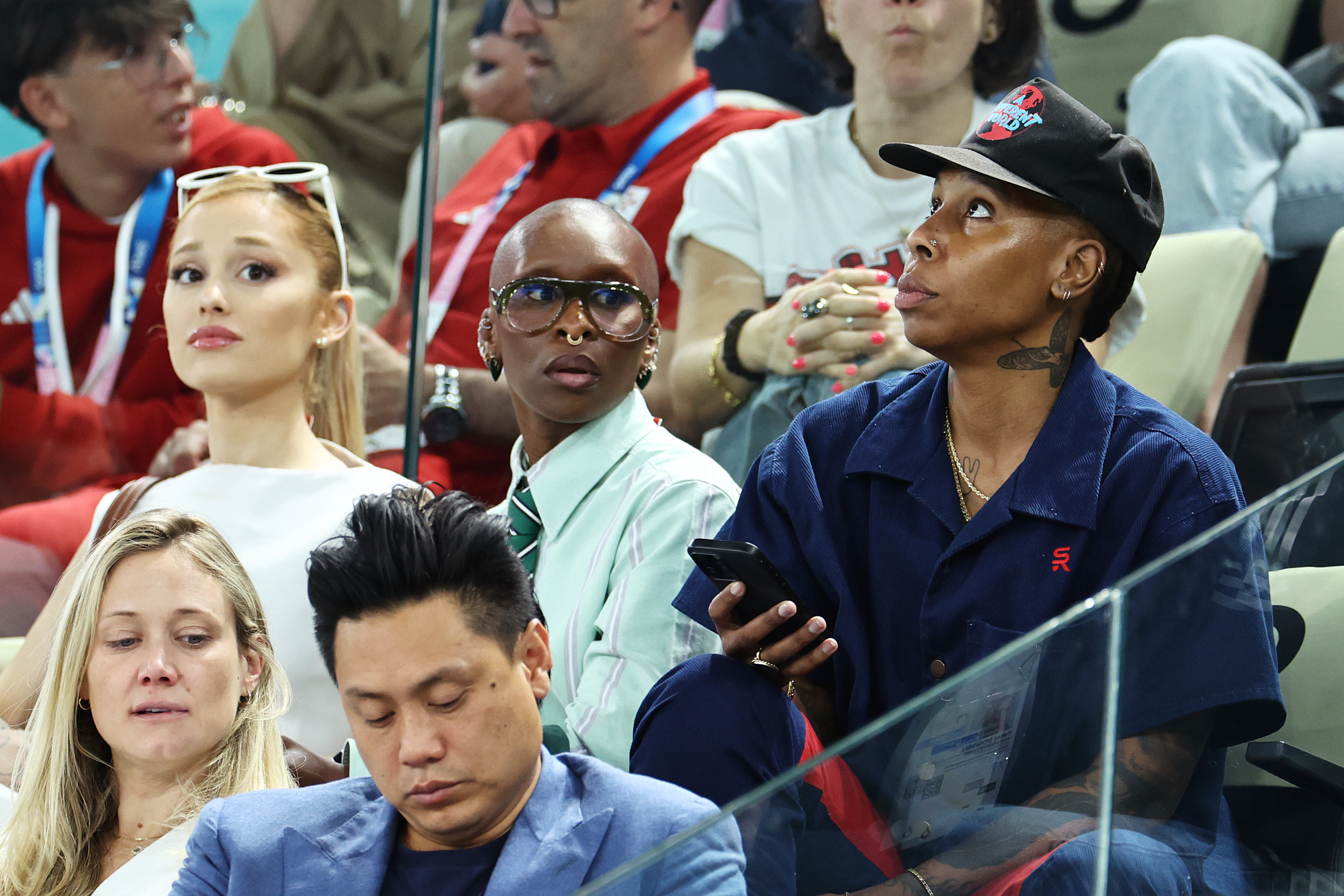 Ariana DeBose, Cynthia Erivo, and Lena Waithe are in a stadium audience. DeBose wears a white top, Erivo a green-striped tie and glasses, and Waithe a navy shirt and cap