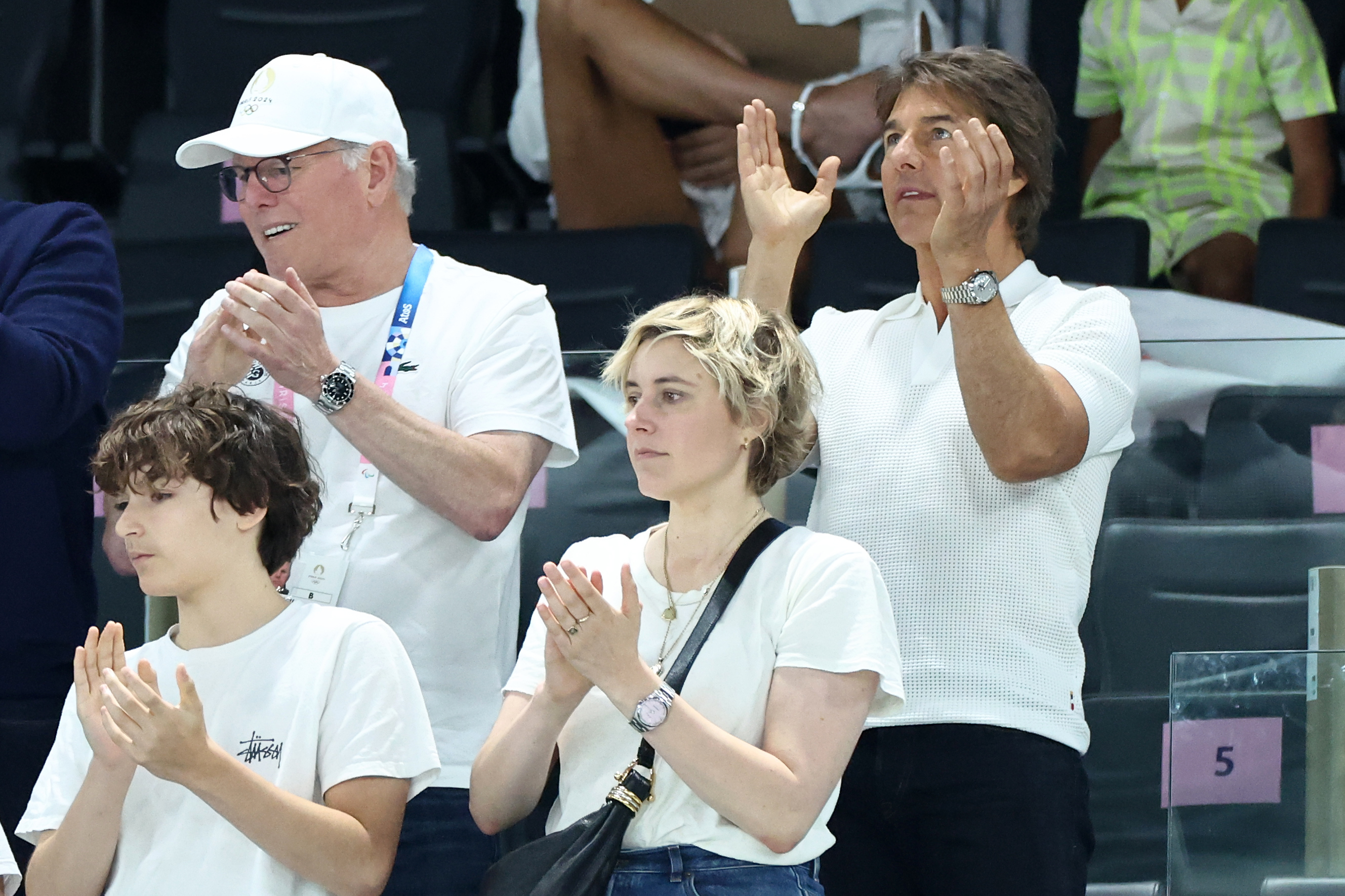 David Zaslav, Rose Gilroy, Tom Cruise, and a child are applauding at a sporting event. Tom and David wear white shirts, and Rose wears a white t-shirt with jeans
