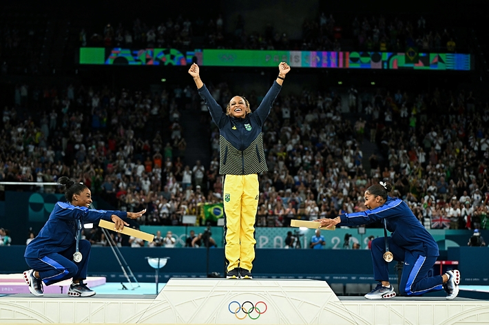 Rebeca Andrade stands in victory on the Olympic podium, flanked by two other gymnasts presenting her with medals