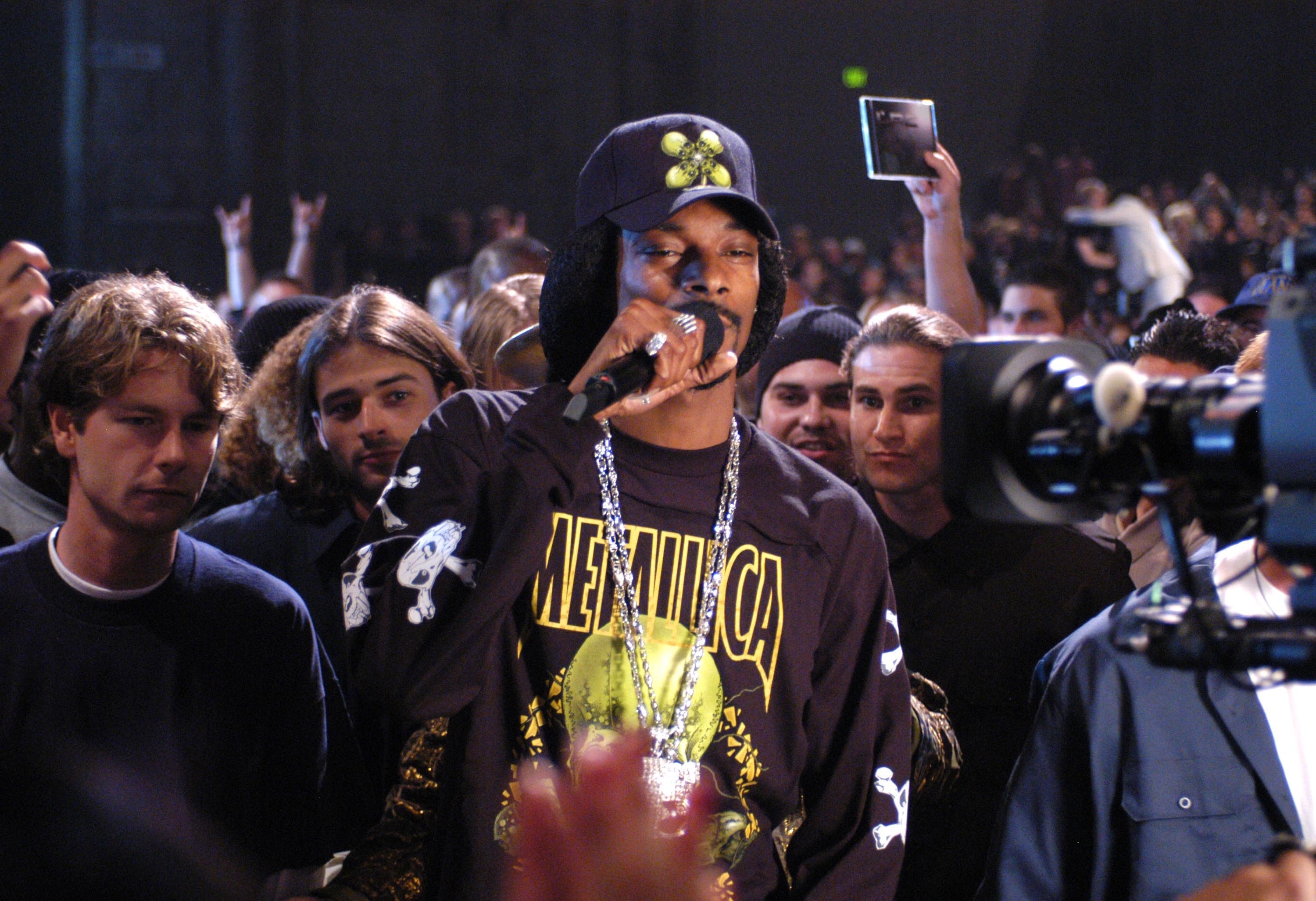 Snoop Dogg wearing a Metallica shirt and a hat with pins, rapping into a microphone at a crowded event. Several unidentified attendees are in the background