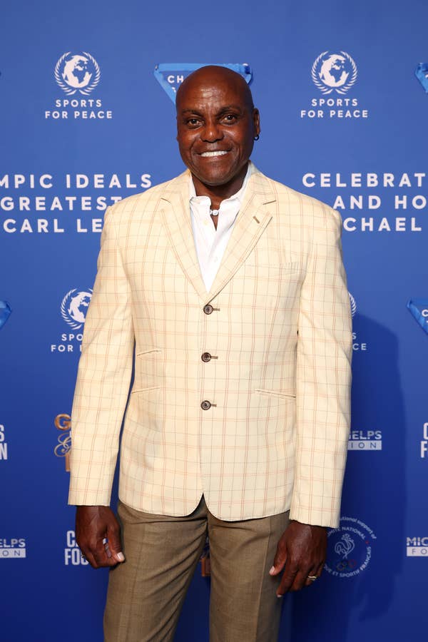 Carl Lewis in a light checkered blazer, white shirt, and brown pants at a Sports for Peace event with a blue backdrop