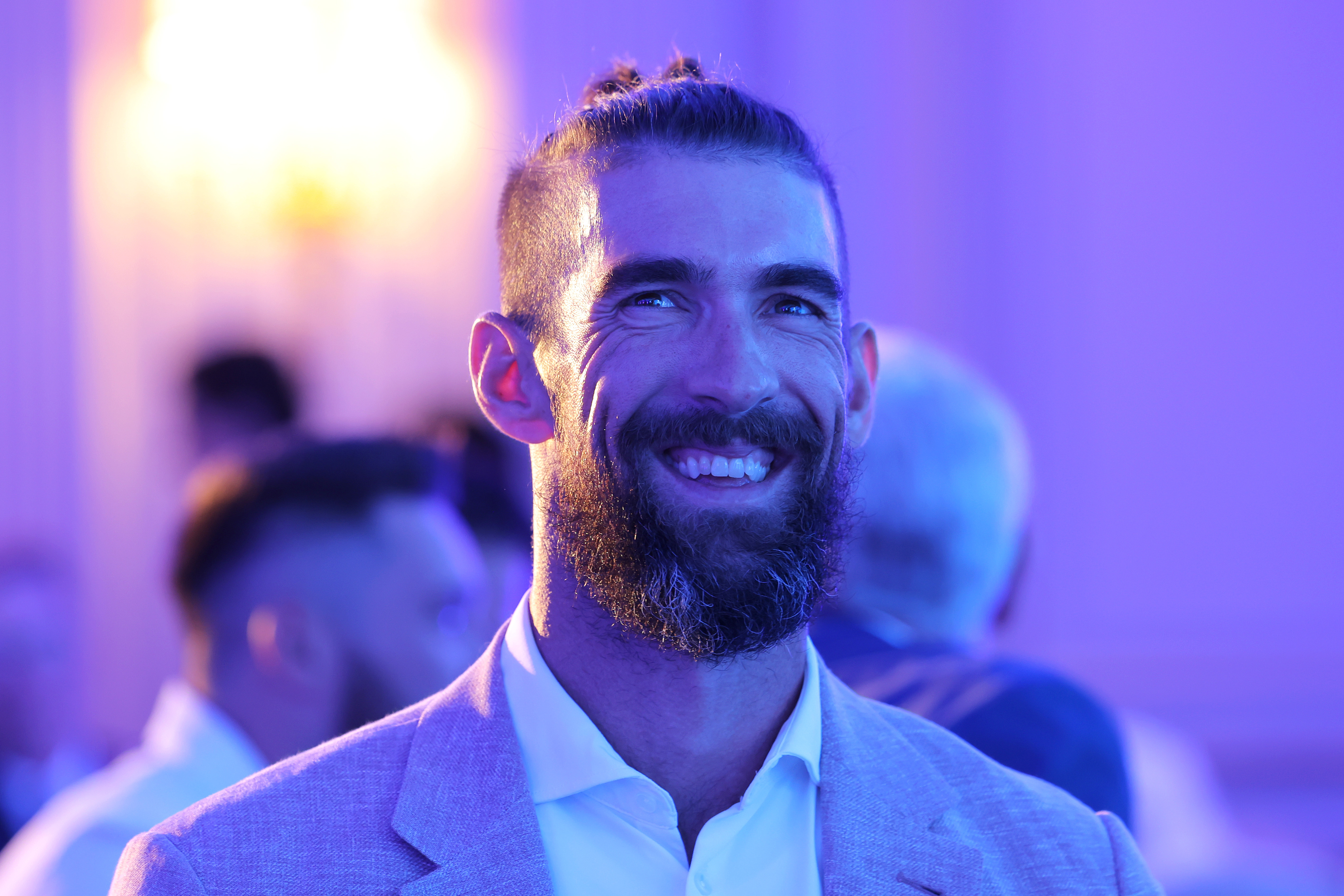Michael Phelps at an event, smiling, wearing a light-colored suit jacket with a man bun hairstyle and beard