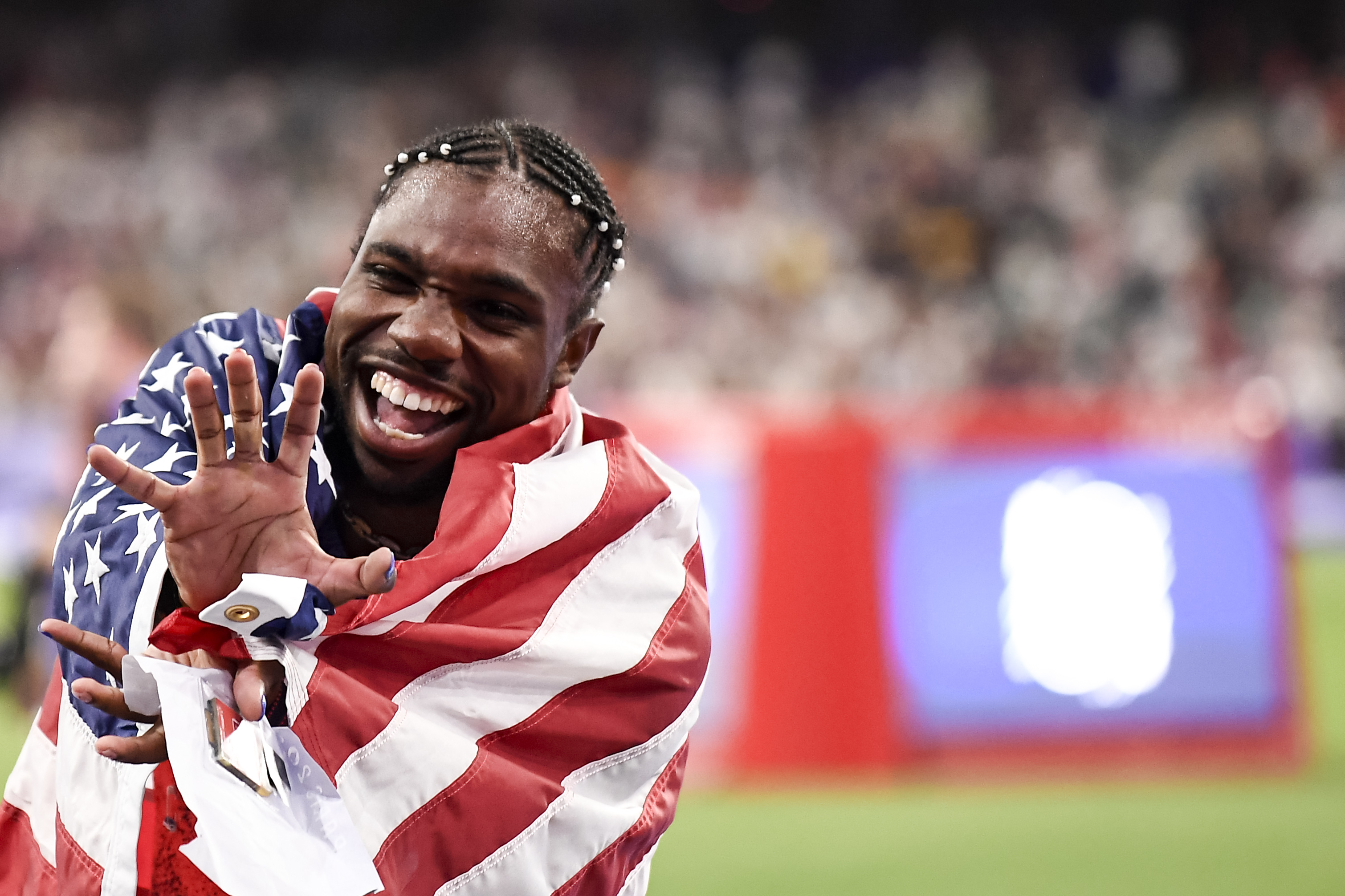 Noah Lyles wraps himself in the American flag and smiles joyfully on a sports field