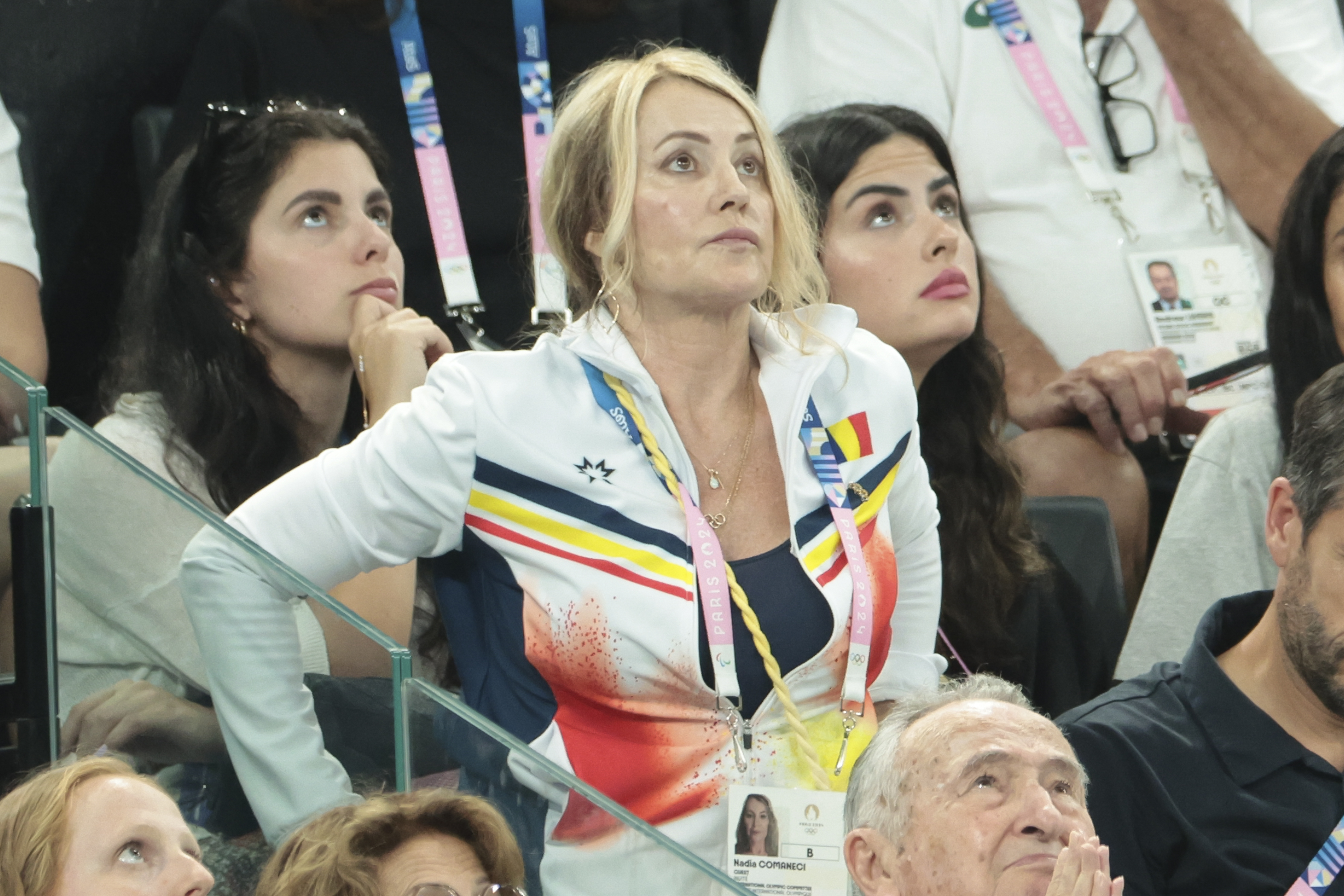 Three women, including Nadia Comăneci, attentively watching an event in a crowded stadium.