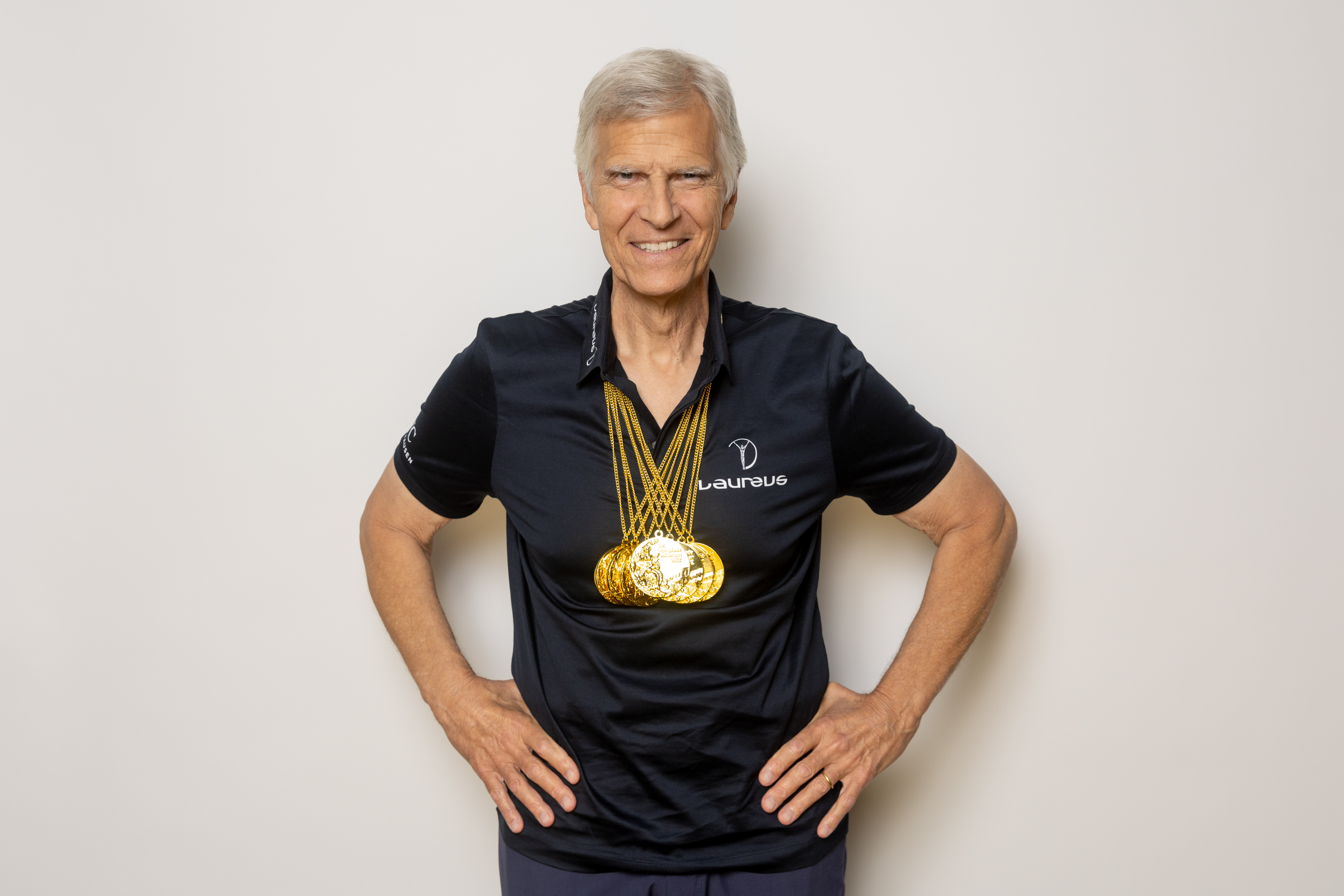 Mark Spitz stands smiling with hands on hips, wearing a black Laureus shirt and several gold medals around his neck