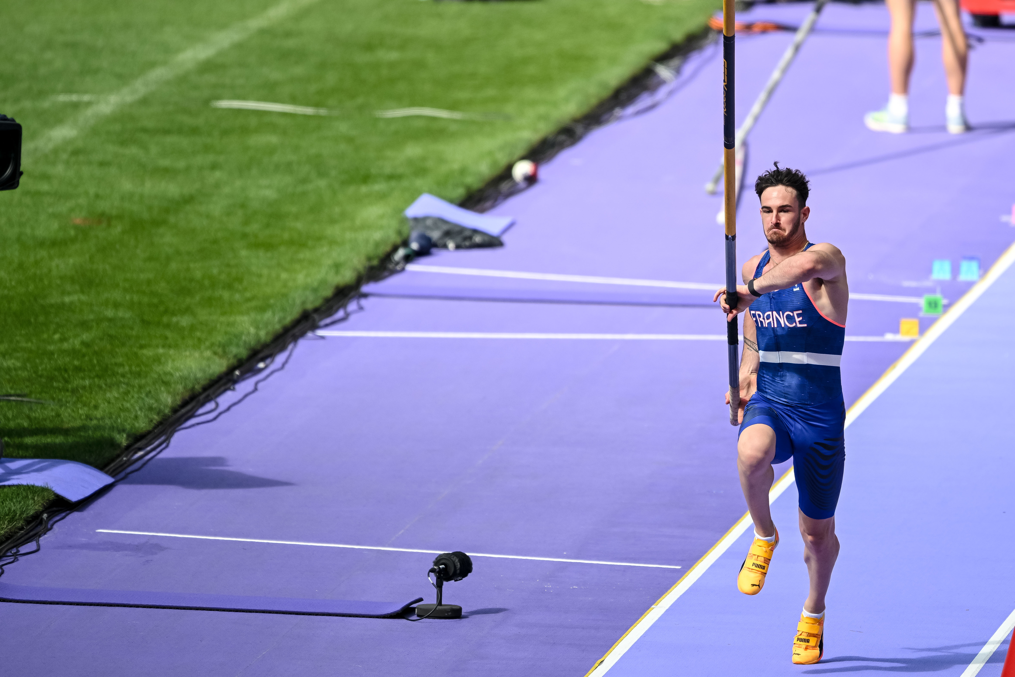 Athlete attempting a pole vault during an event, with a focus on the run-up phase. The athlete is wearing a sports uniform that says &quot;France.&quot;