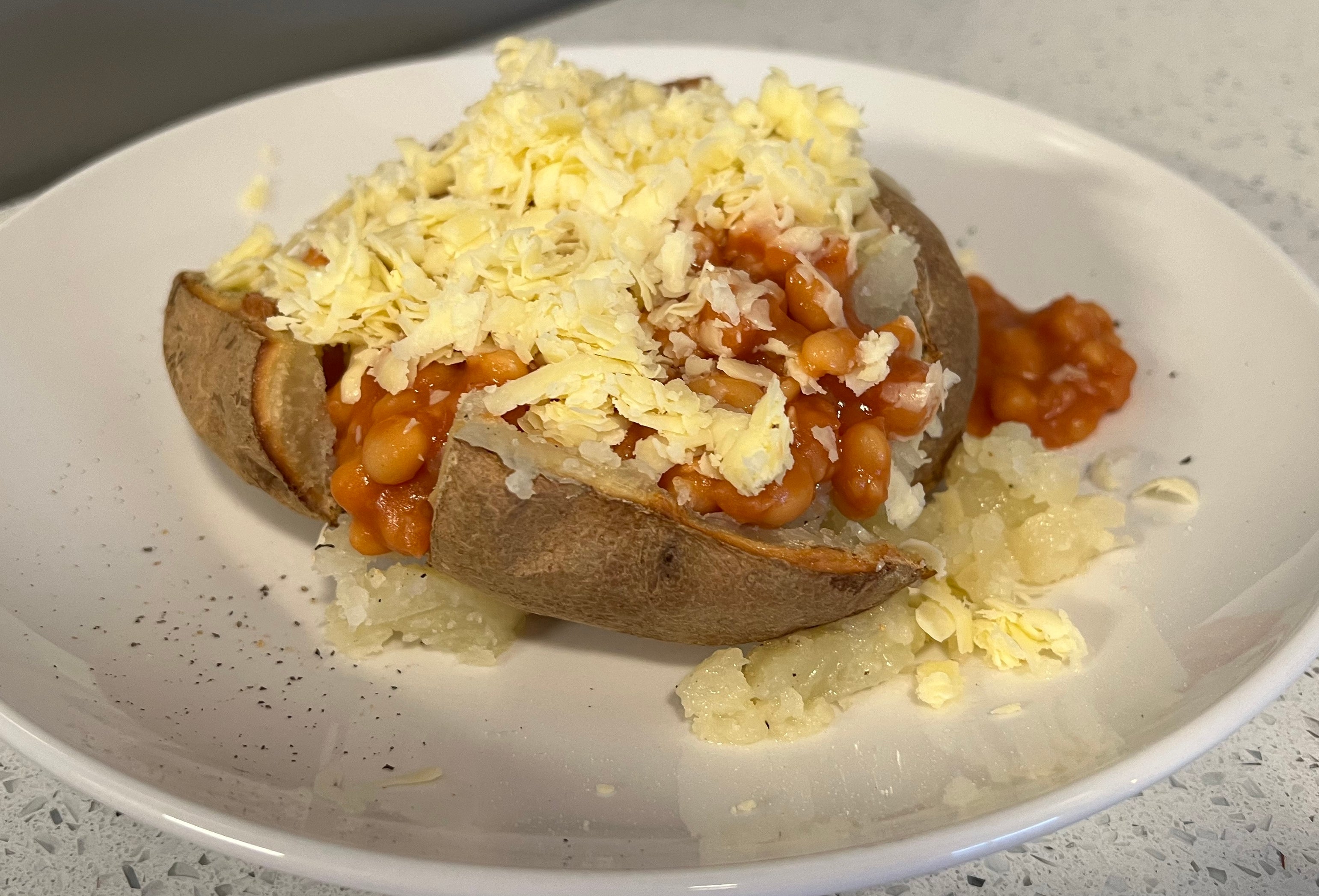 A baked potato topped with baked beans, grated cheese, and a sprinkle of black pepper, served on a white plate