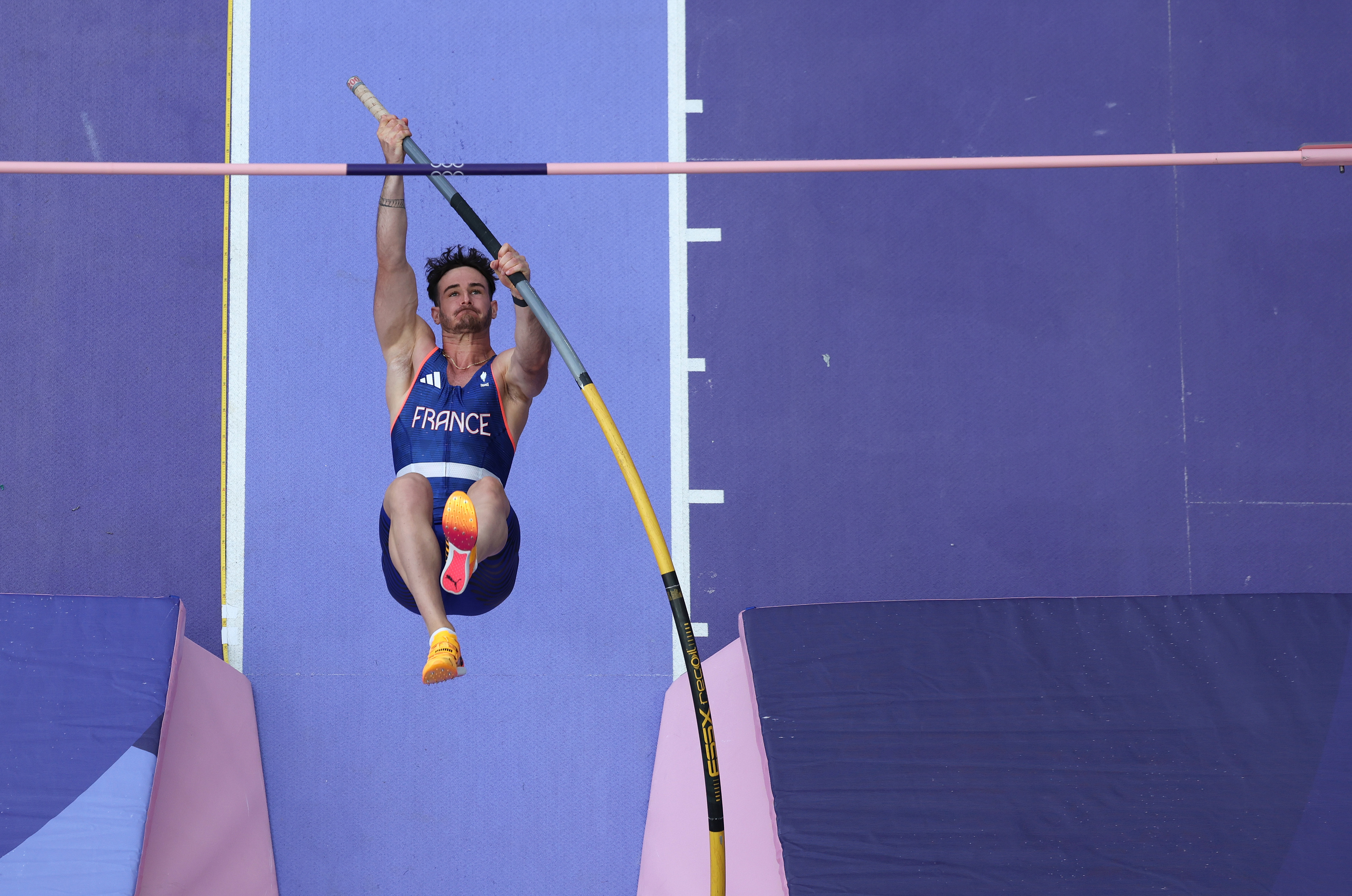 A male athlete in a &quot;France&quot; uniform is pole vaulting, extending his body upwards with the pole in mid-bend during a competition