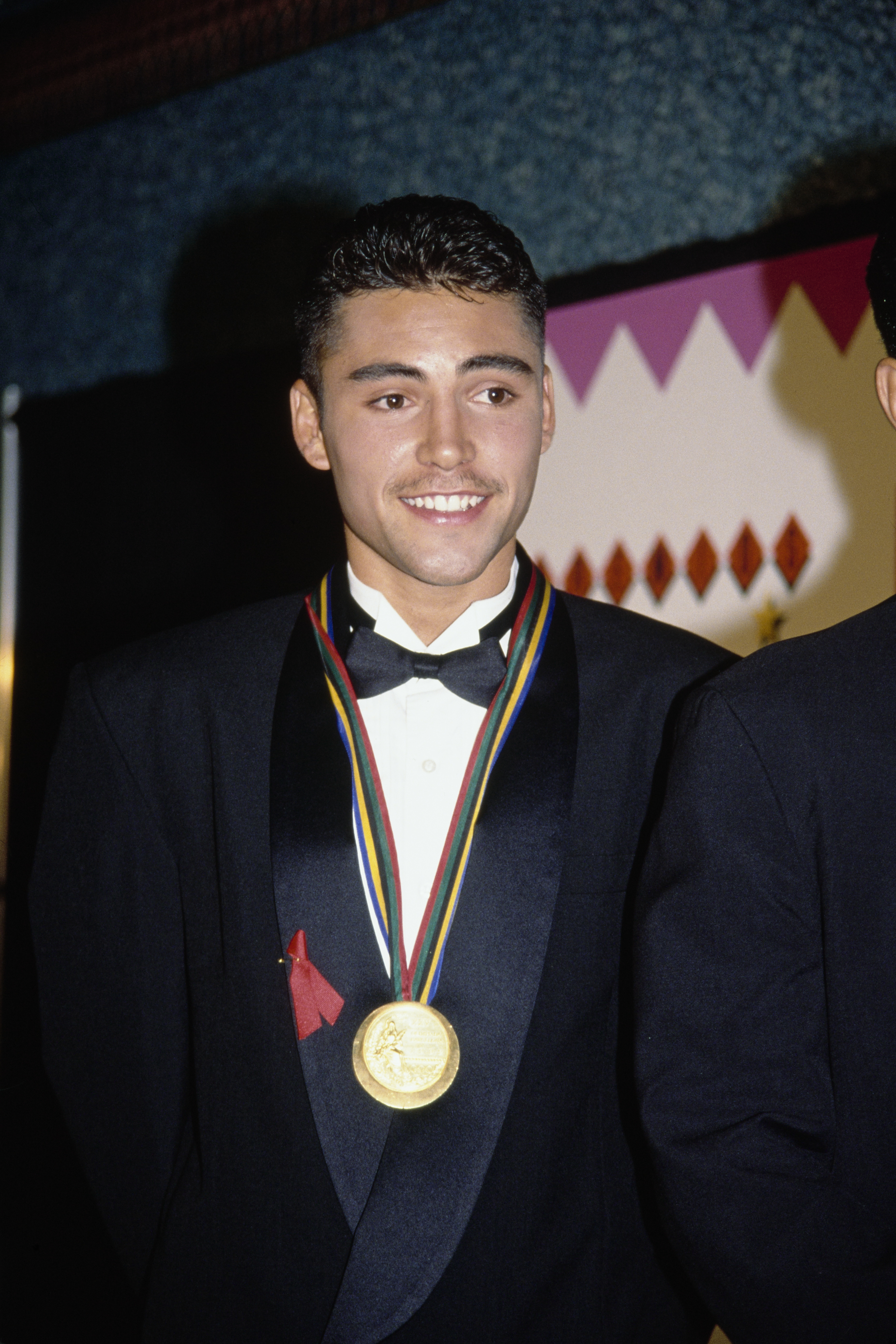 Oscar De La Hoya, wearing a tuxedo and gold medal, smiles at an event