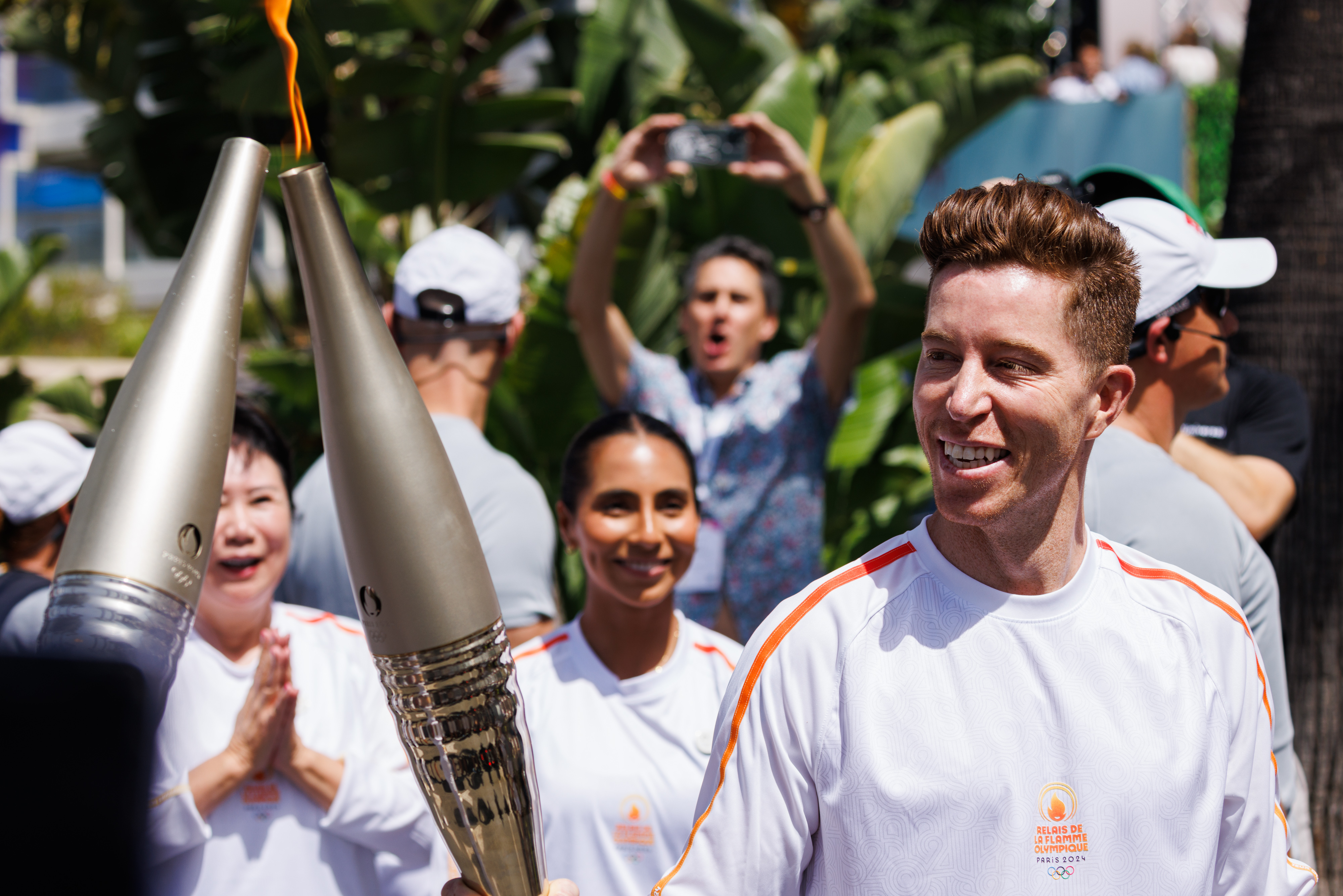 Shaun White, holding a torch, smiles while participating in a relay event with other runners, who also carry torches. People cheer in the background