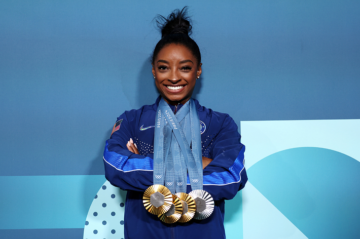 Simone Biles smiling and posing with five Olympic medals around her neck, wearing a team jacket