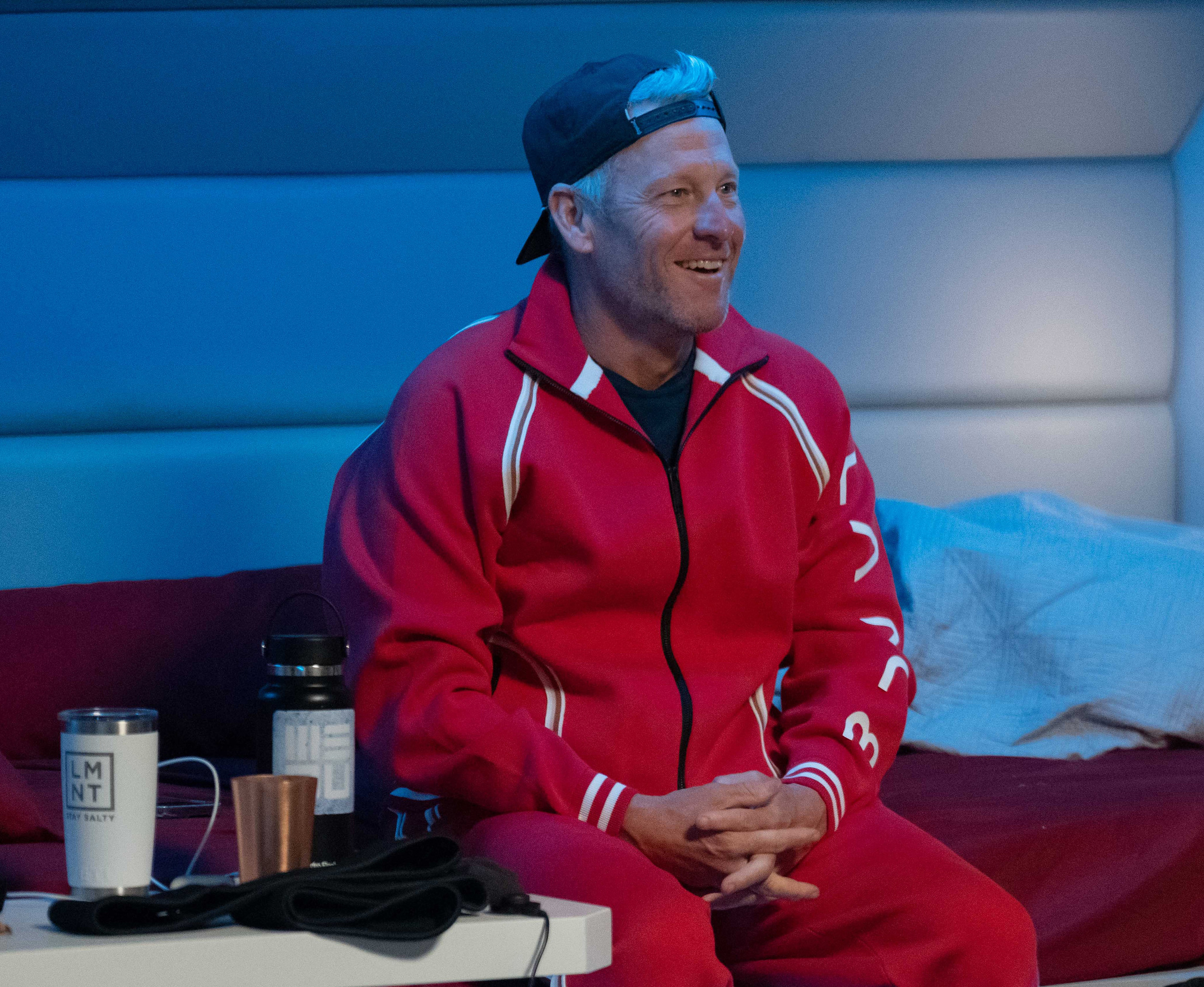 Lance Armstrong sits on a bed, wearing a casual tracksuit and a backward cap. Nearby, there are drinks on a side table
