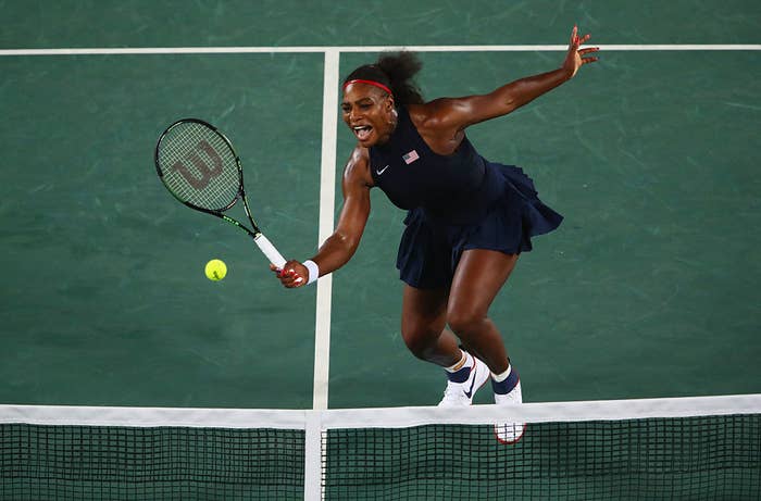 Serena Williams lunging to hit a tennis ball with her racket during a match on a tennis court. She is wearing a sleeveless athletic dress and sneakers