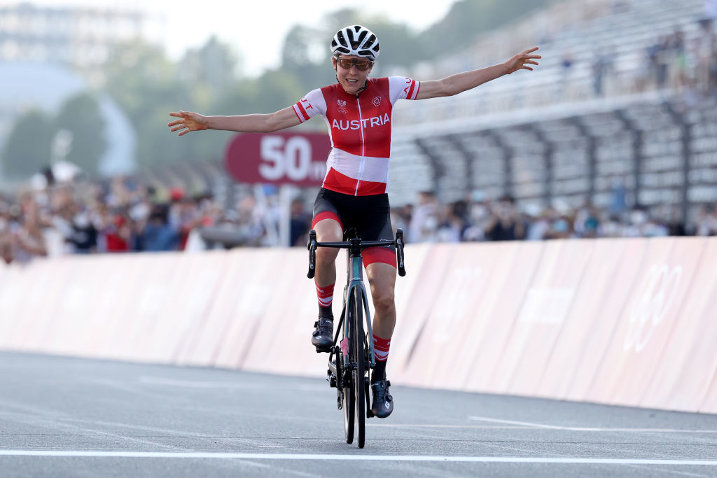 Anna celebrates crossing the finish line with arms outstretched at a race track, with spectators in the background
