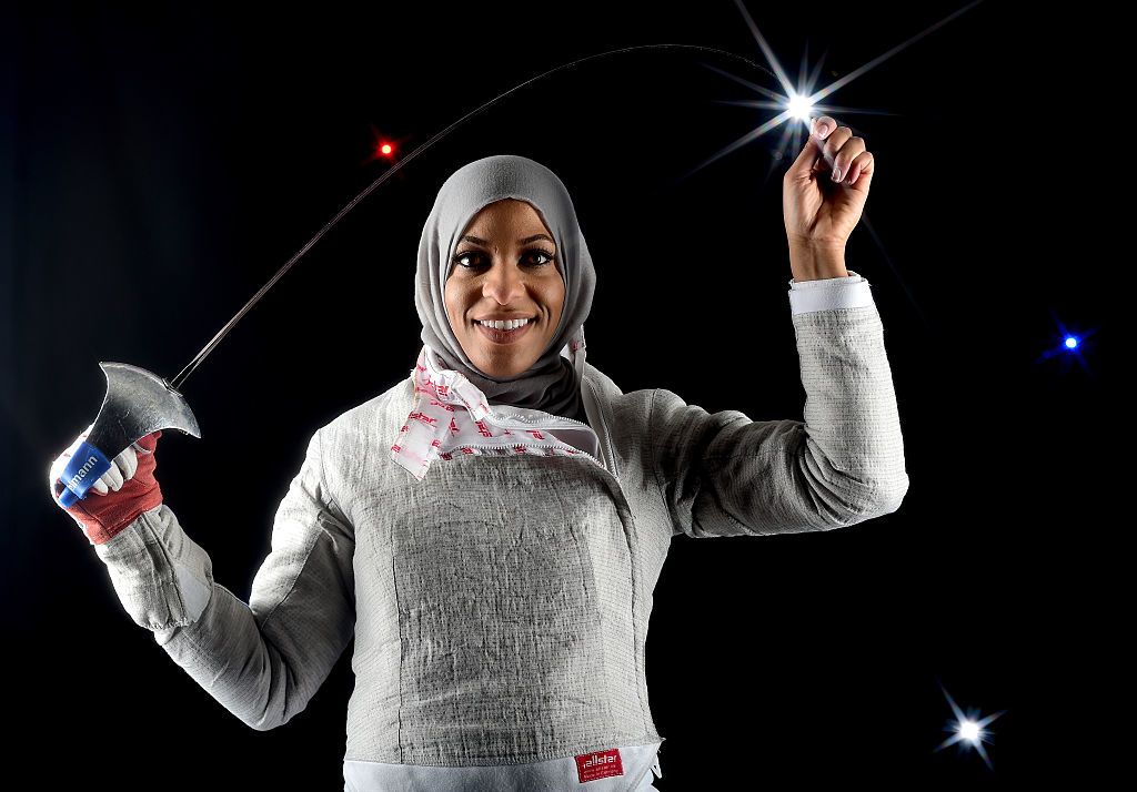 Ibtihaj Muhammad, wearing fencing gear and holding a foil in one hand, poses confidently with her medal gleaming