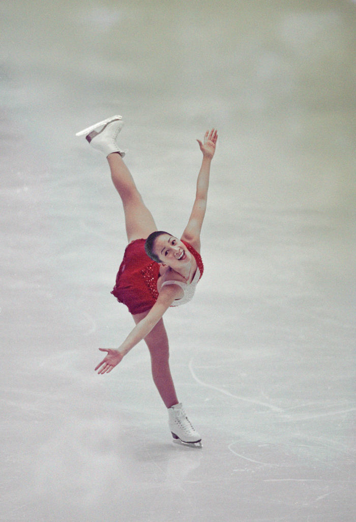 Michelle in mid-performance, wearing a sparkling costume with a skirt. Her leg is extended high behind, arms outstretched, and a smile on her face