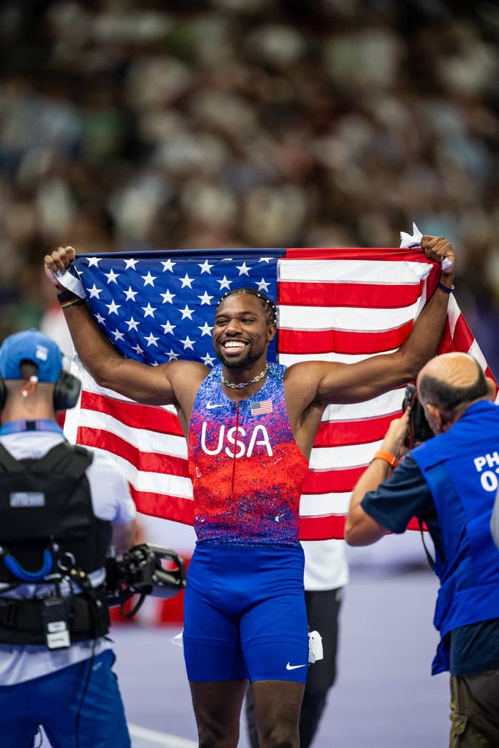 Noah Lyles holds the USA flag, celebrating his victory. He wears an athletic outfit with "USA" printed on the chest. Photographers capture the moment