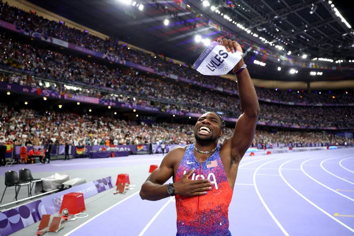 Noah Lyles celebrates victory on the track, holding a name placard overhead, surrounded by a cheering crowd in a stadium