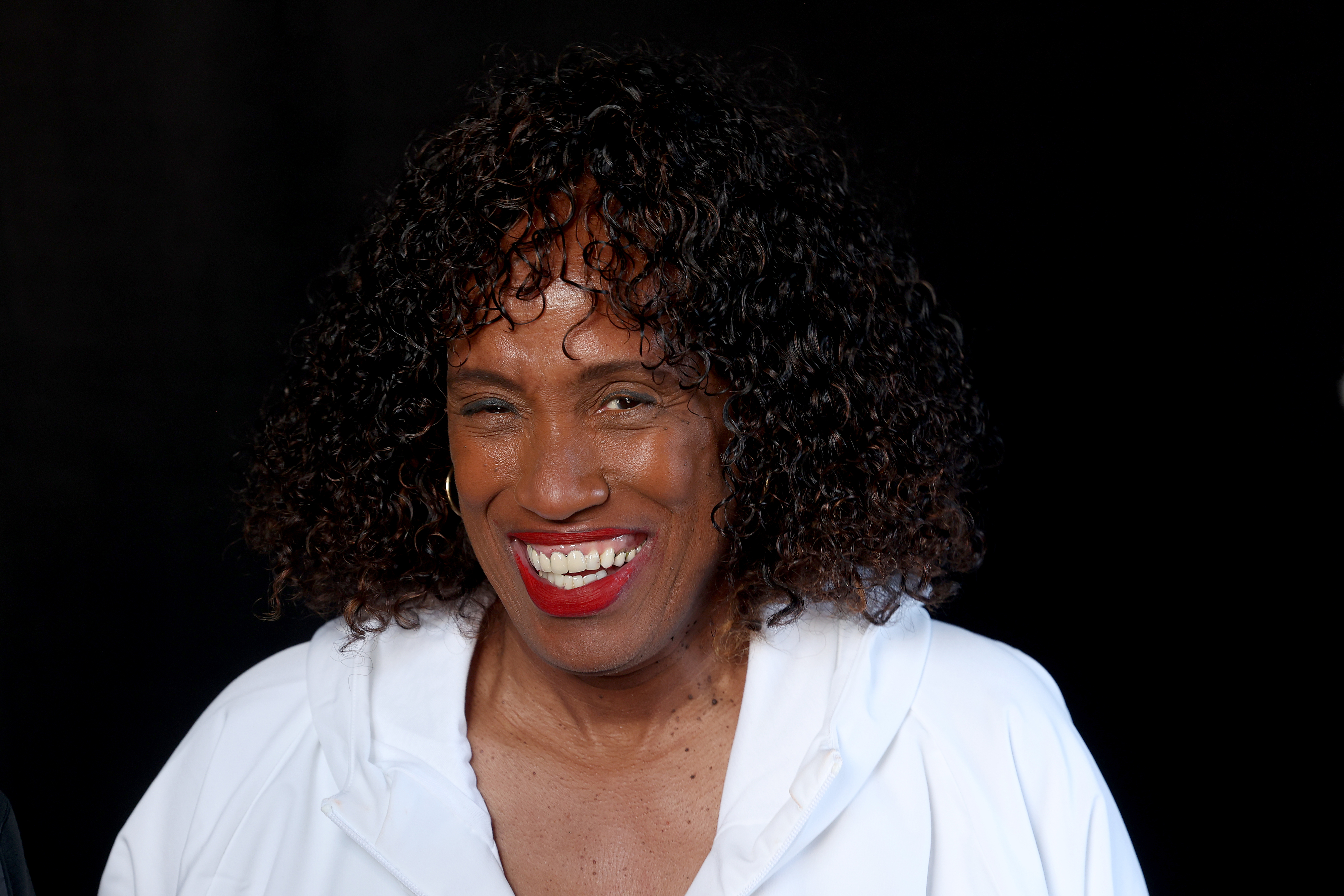 Jackie Joyner-Kersee with curly hair wears a white hoodie against a dark background. Names of people in the image are unknown