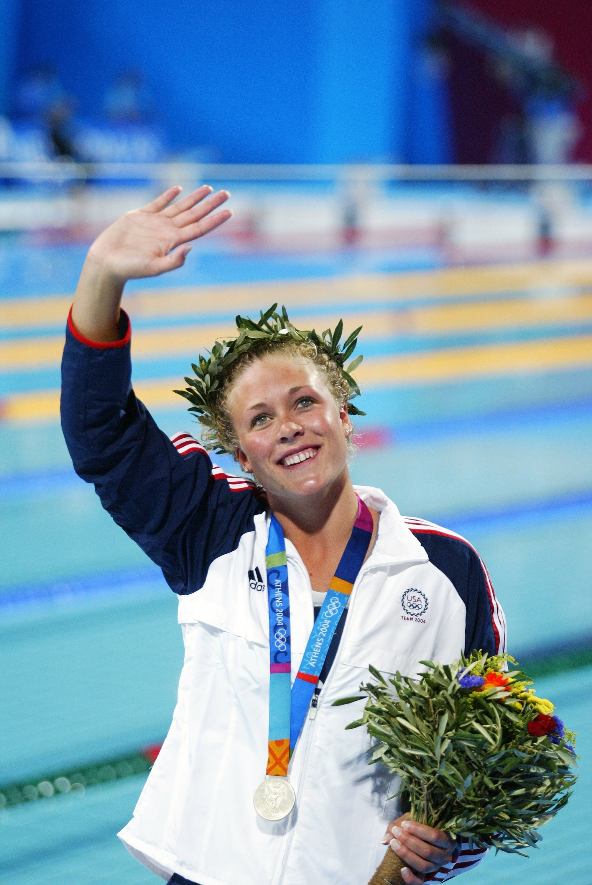 enny Thompson wearing an Olympic medal and a laurel wreath waves while holding a bouquet of flowers at a swimming pool
