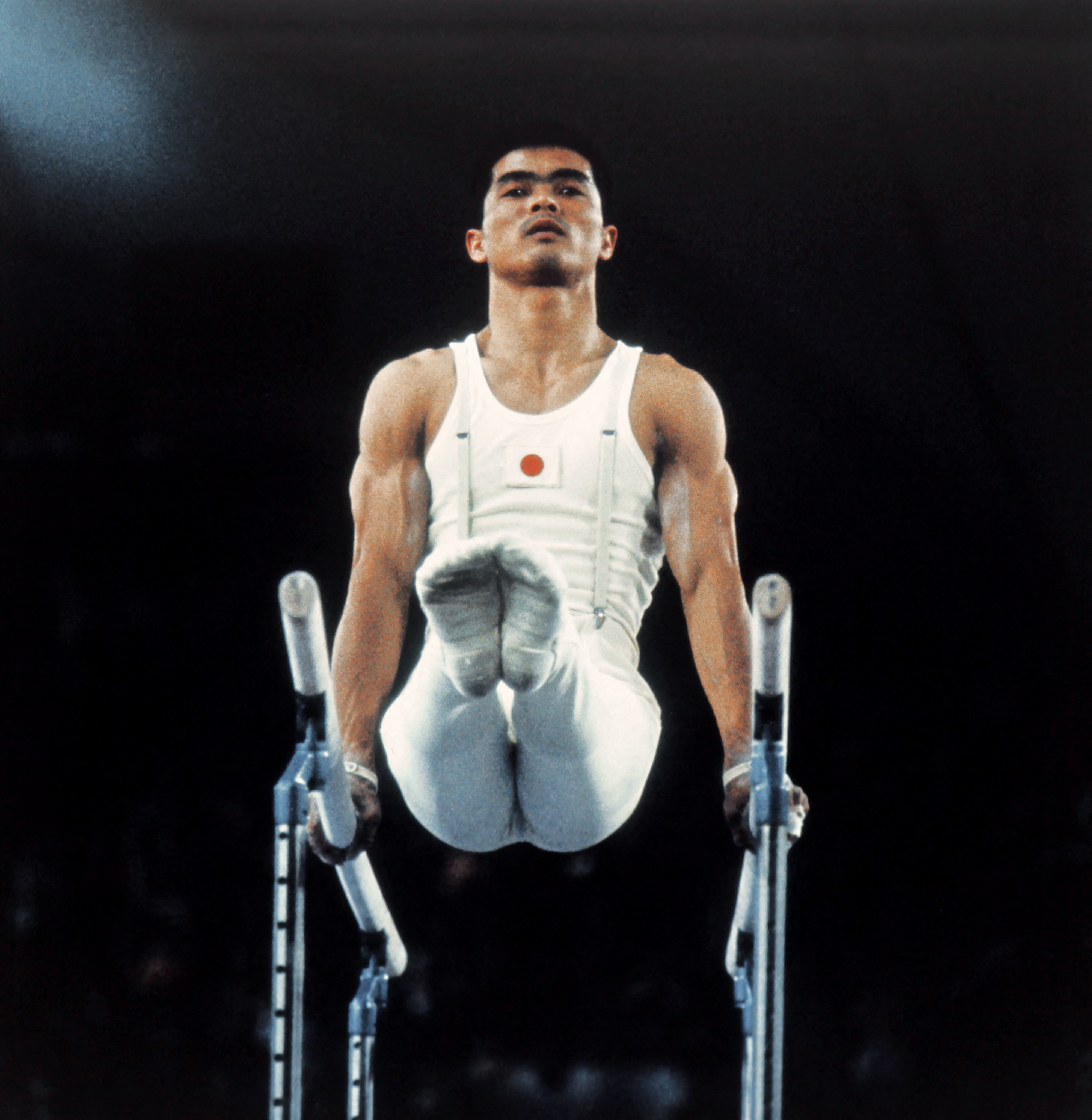 Sawao Kato performing a precision move on parallel bars, wearing a white leotard featuring a red circle