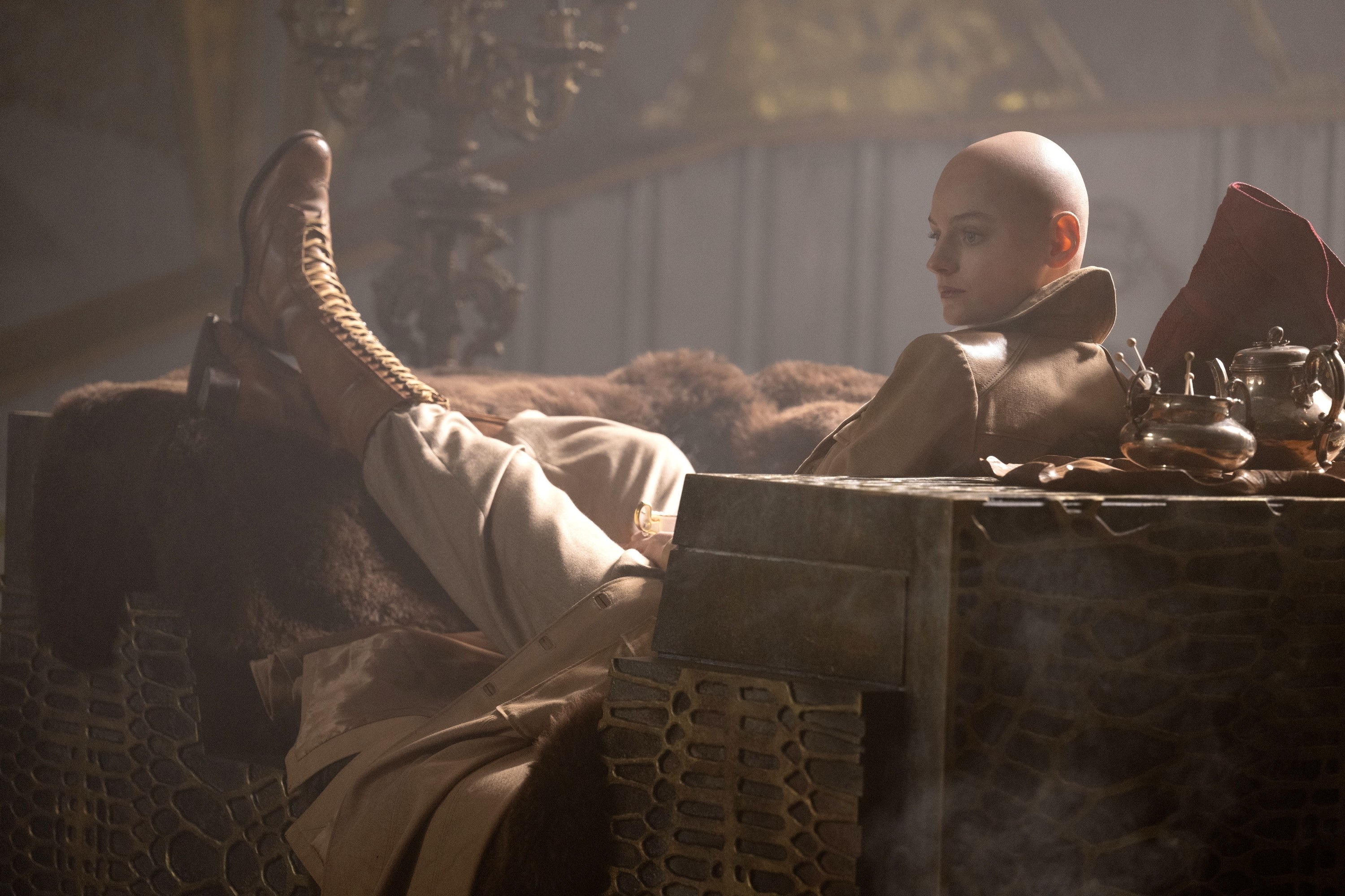 Tilda Swinton lounges on a set in a leather coat and high boots, portraying a character with a shaved head in a scene from a movie