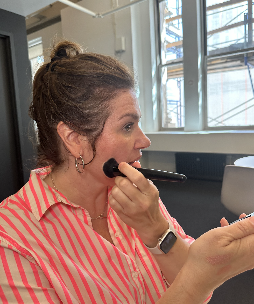 A woman in a pink-striped shirt applies makeup using a brush while looking into a mirror