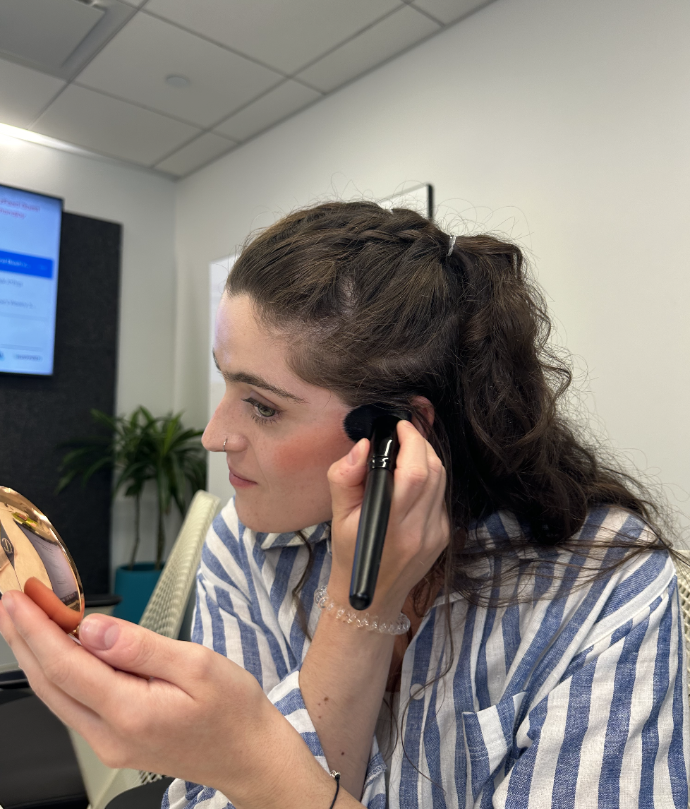 Woman in a striped shirt applies makeup with a brush while looking into a mirror