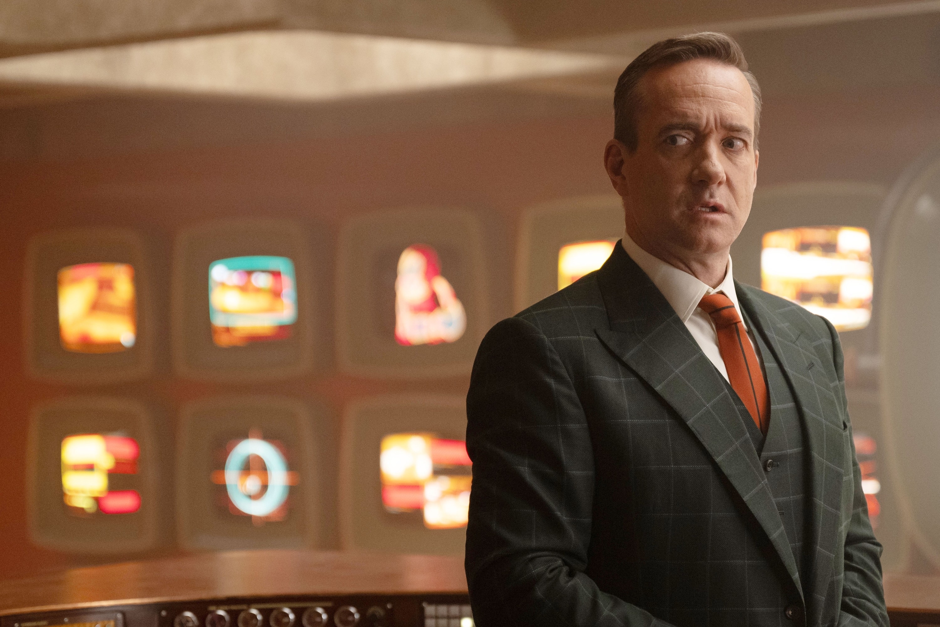 Matthew Macfadyen stands in a control room with multiple vintage screens behind him, wearing a dark checkered suit and an orange tie