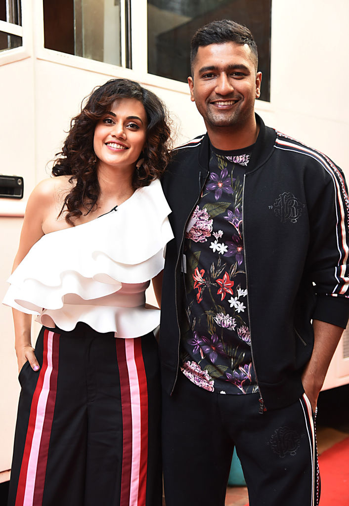 Taapsee Pannu and Vicky Kaushal pose together. Taapsee wears a one-shoulder ruffled top and striped pants. Vicky wears a floral shirt and a track jacket