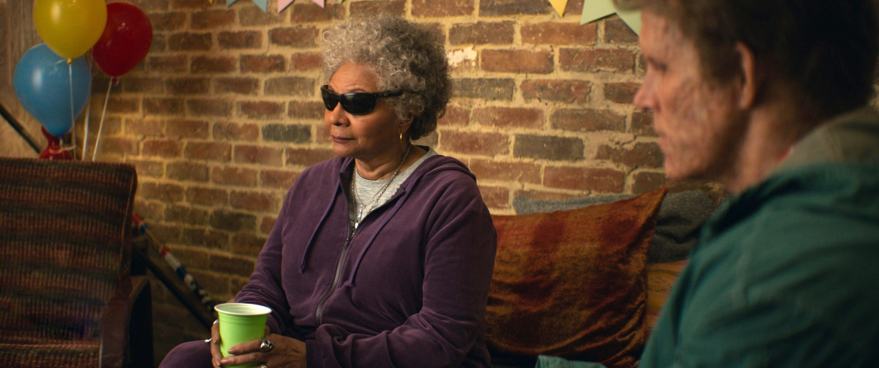 An elderly woman wearing sunglasses and a purple tracksuit holding a green cup sits indoors with an elderly man in green jacket