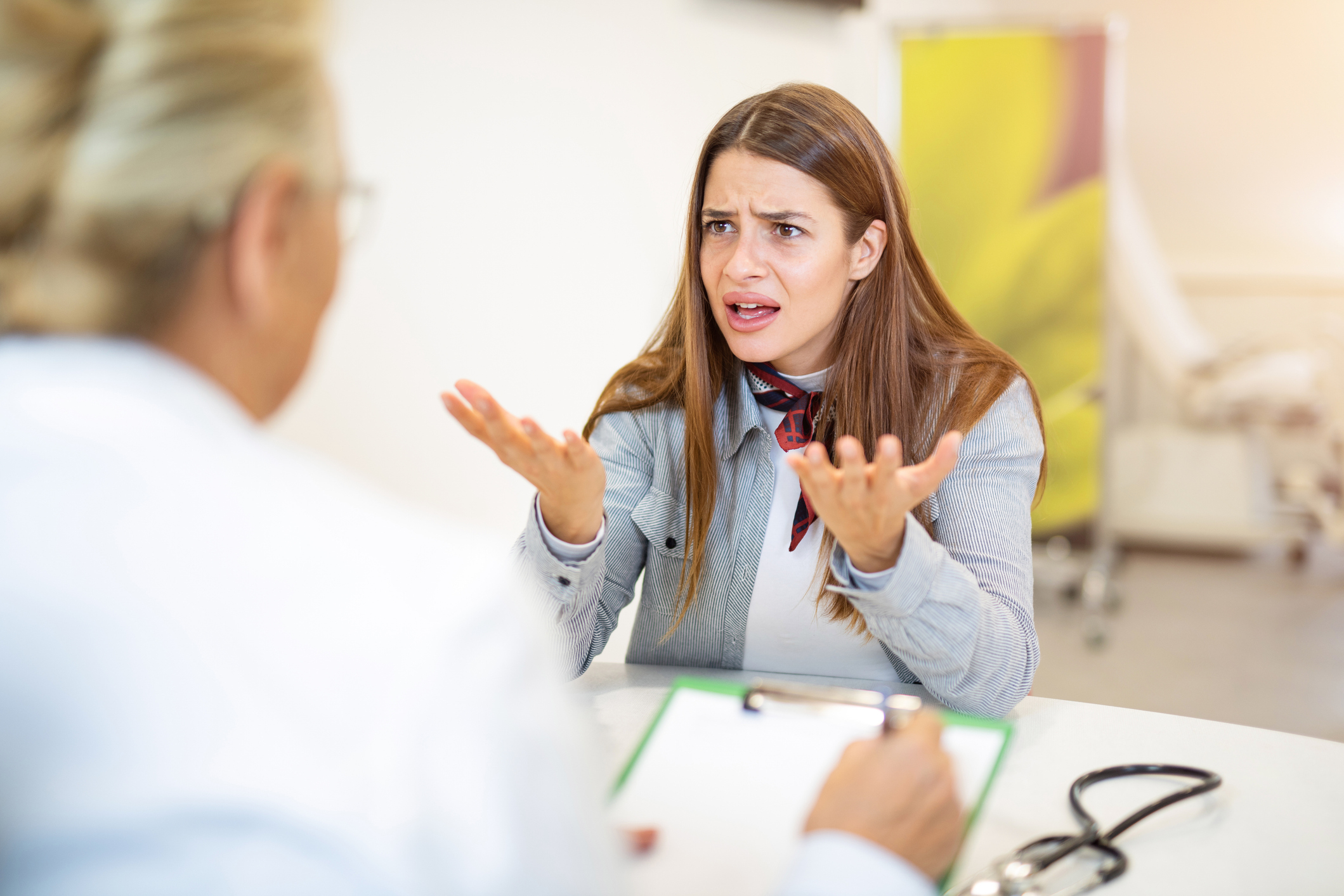 A woman gestures expressively with her hands while speaking to a person holding a clipboard, possibly during a consultation
