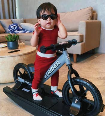A toddler stands confidently connected  a stationary Strider bicycle successful  a surviving  room, wearing sunglasses, a sleeveless outfit, and shoes with reddish  accents