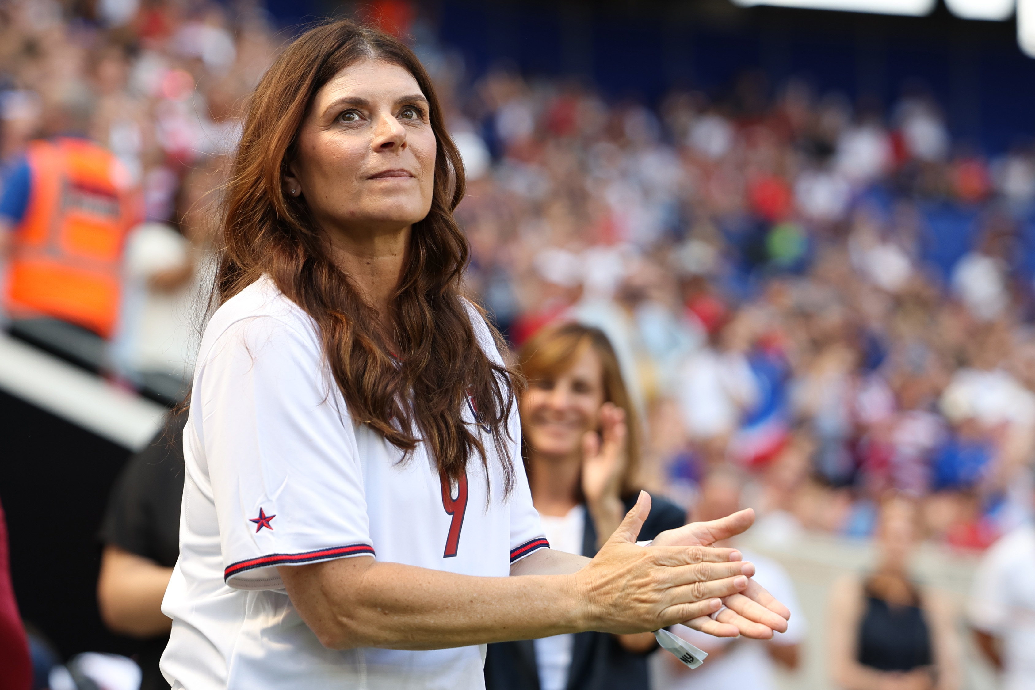 Mia Hamm in a sports stadium, wearing a number 9 jersey, is clapping with the crowd visible in the background