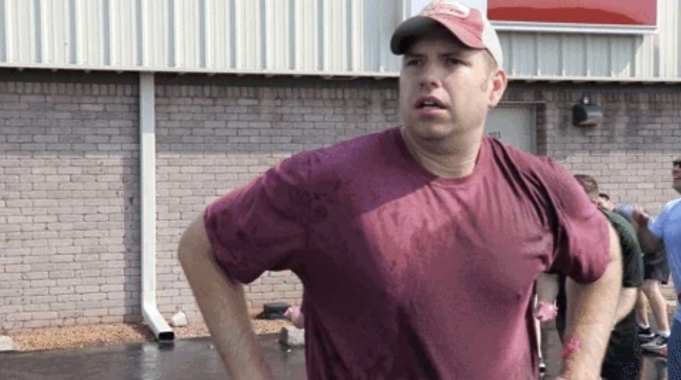 A man in a maroon t-shirt with his hands on his hips stands outside a building. He is wet, possibly from a water activity. Other people are seen in the background