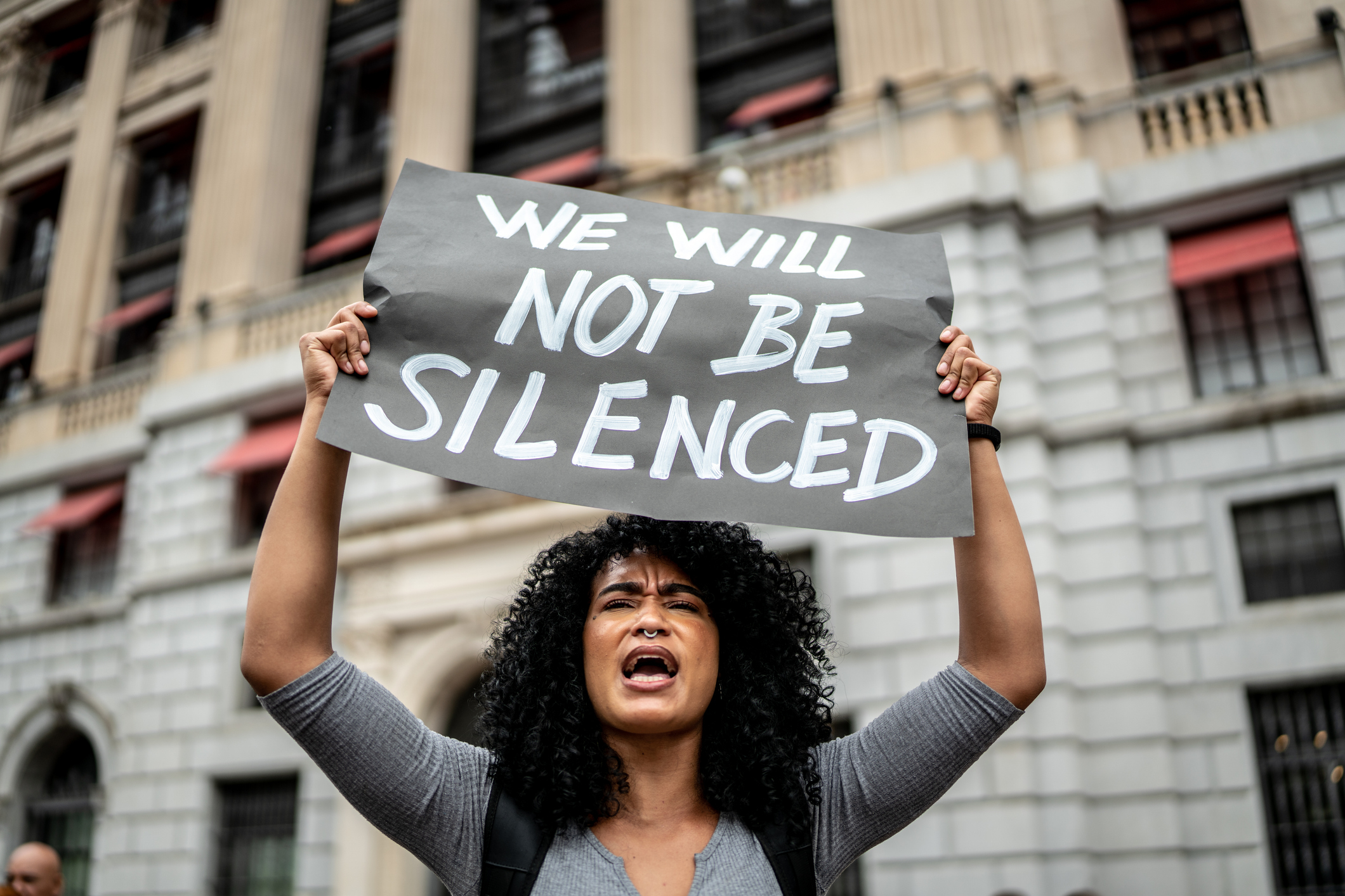 A person holds a sign reading "We will not be silenced" during a protest. They are passionately shouting. The background features a building