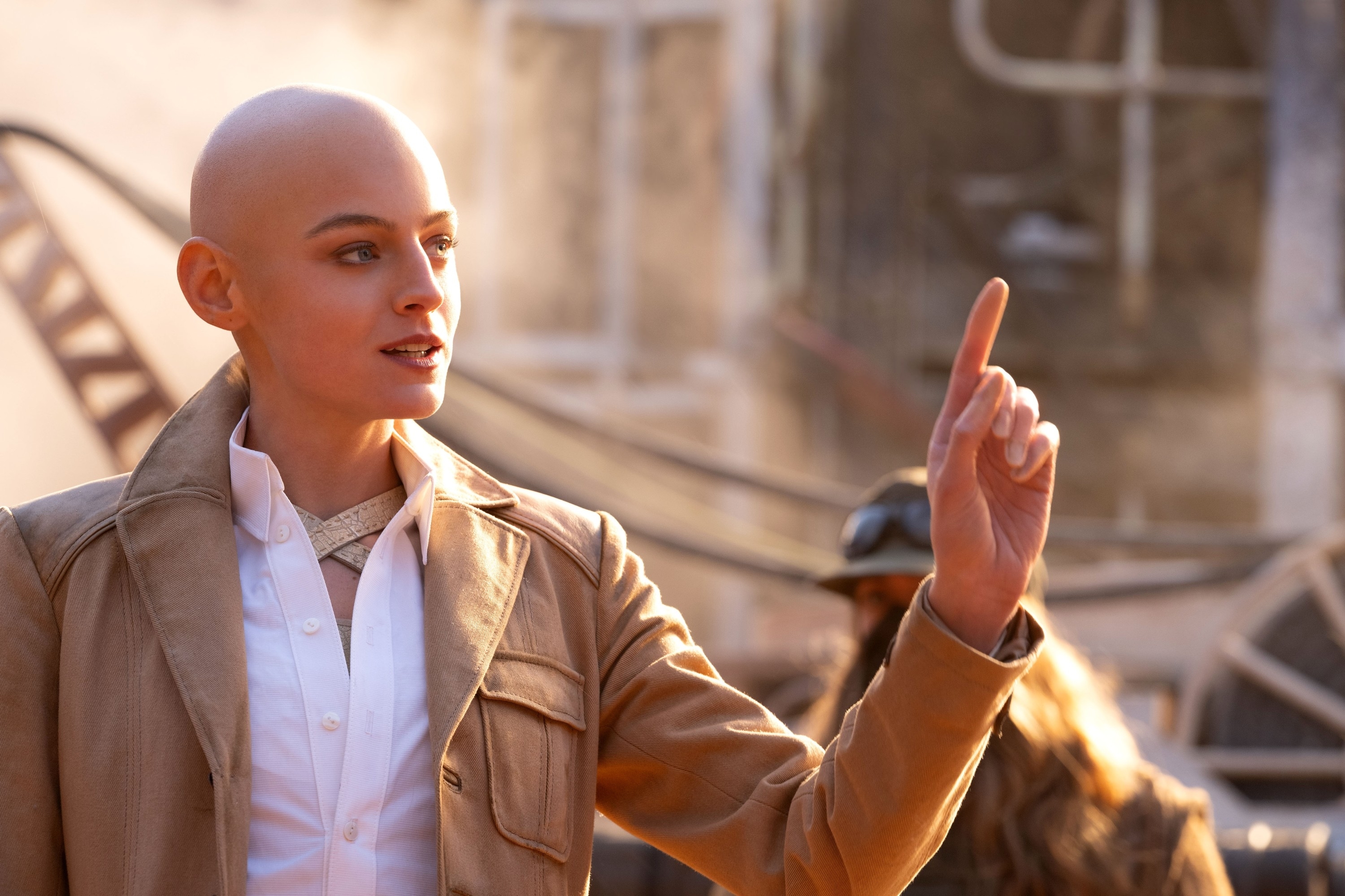 Anya Taylor-Joy as Furiosa from Mad Max prequel wears a beige trench coat and white shirt, gesturing with her finger while speaking in a post-apocalyptic setting