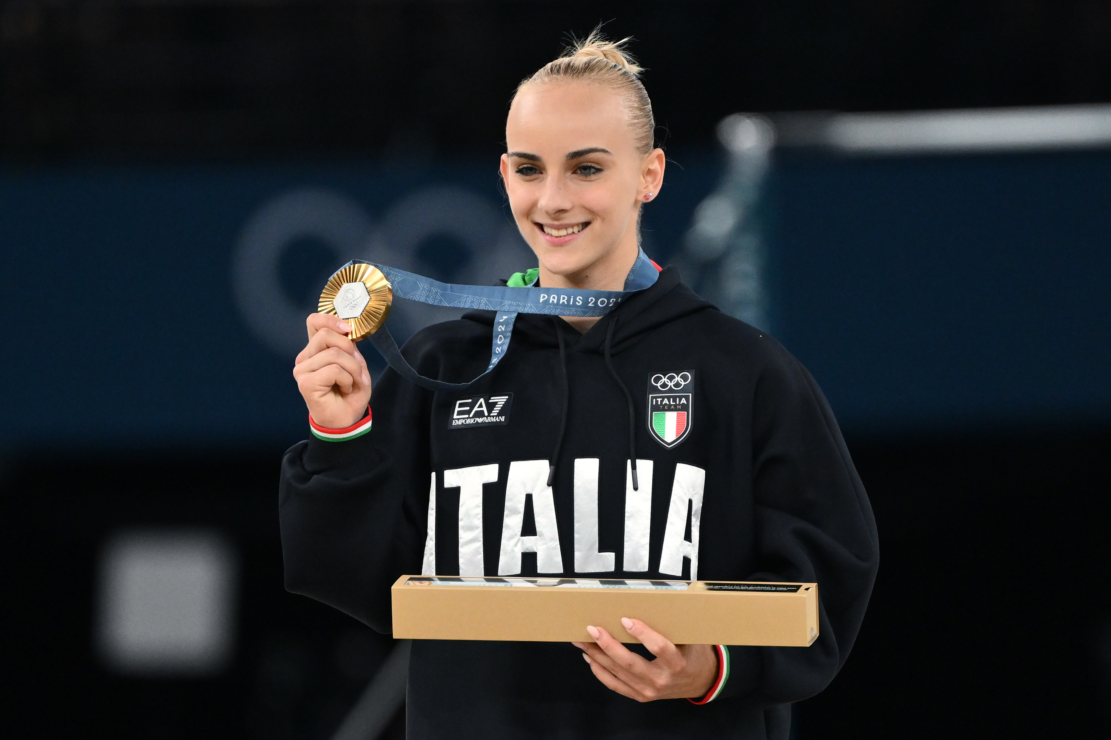 Italian gymnast Giorgia Villa holds up her gold medal in one hand and a certificate in the other, wearing an "Italia" hoodie at a competition event
