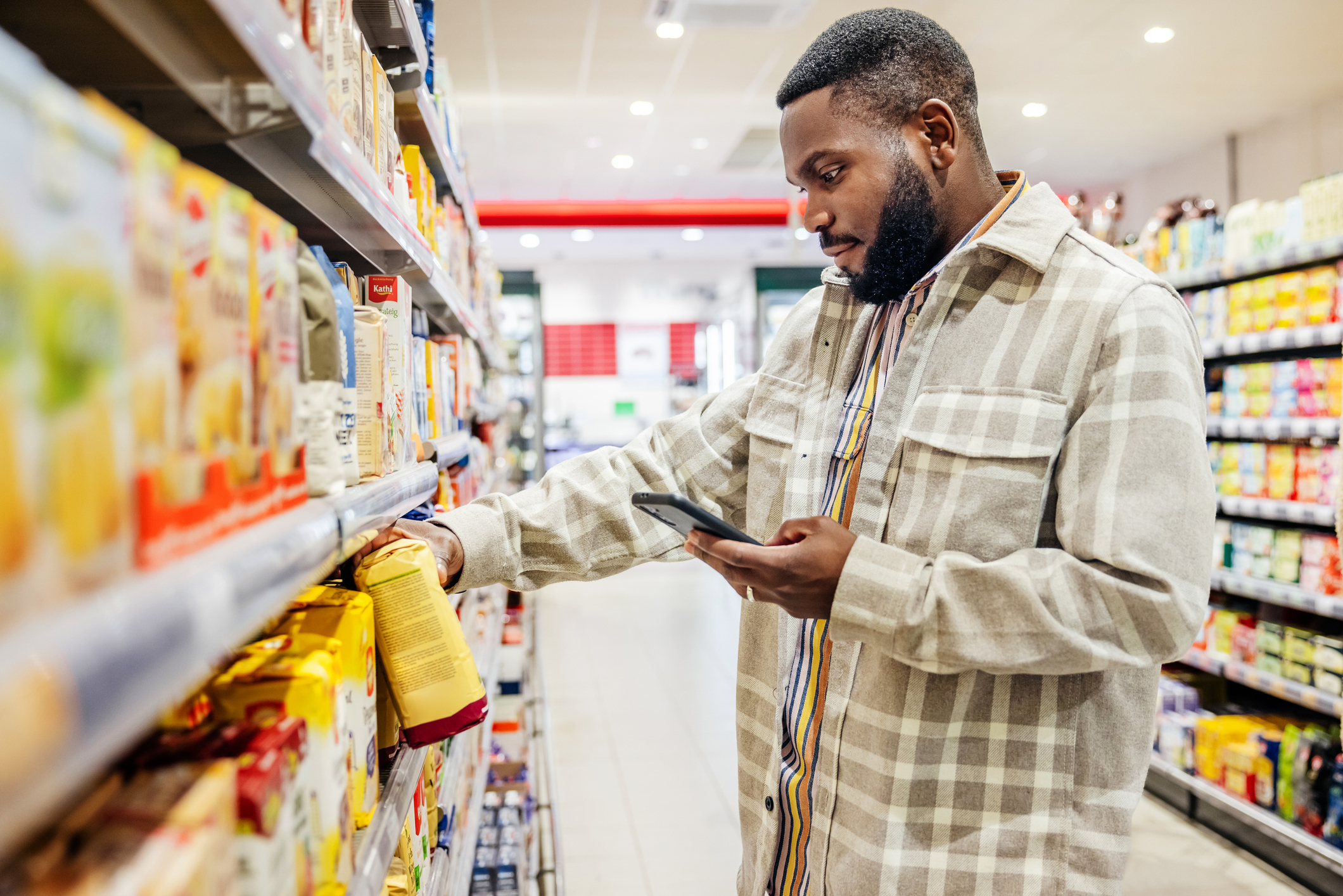 A man in a plaid shirt shops in a grocery store aisle, holding a yellow package in one hand and a smartphone in the other