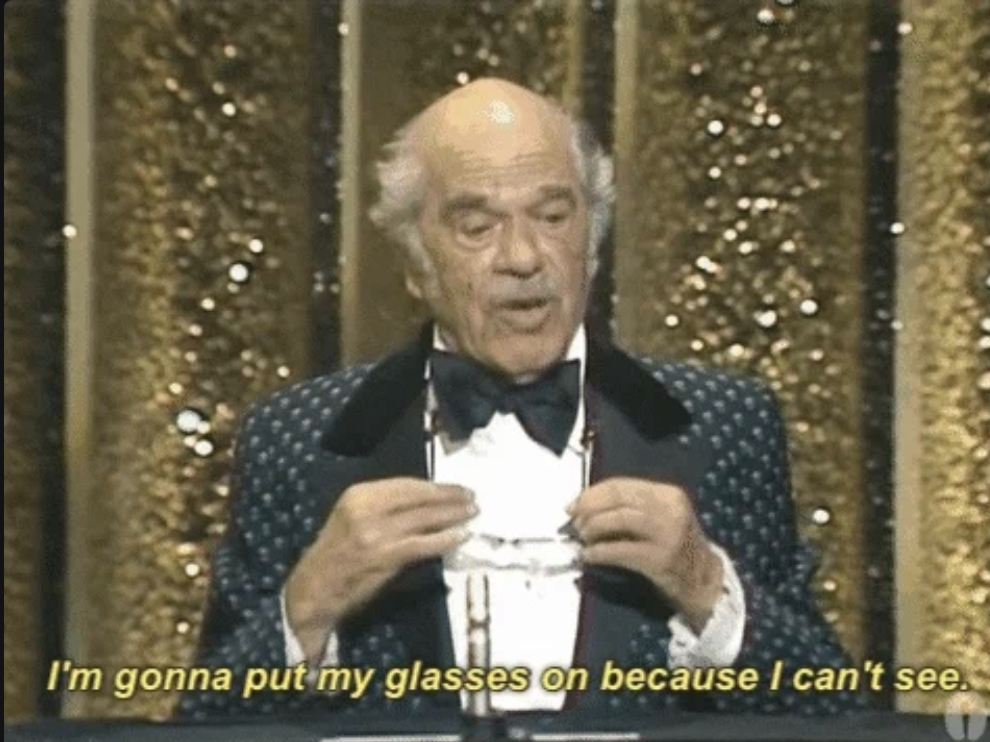 Elderly man in a tuxedo holding glasses near his face, with text, "I'm gonna put my glasses on because I can't see."