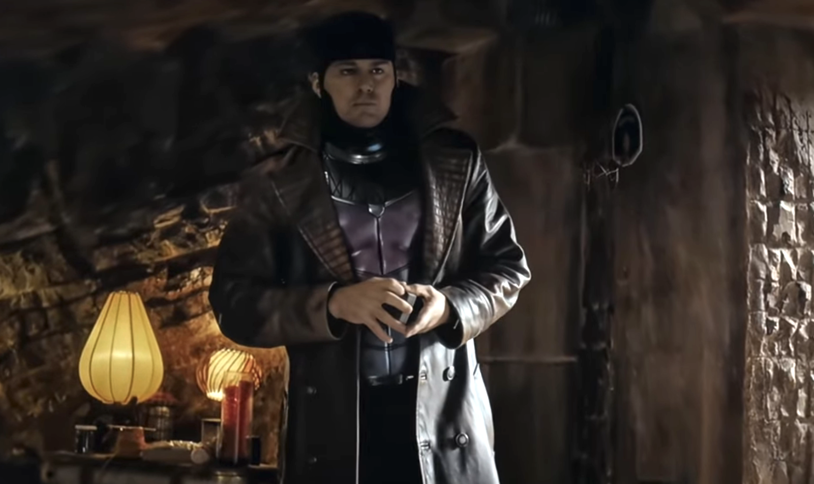 Pedro Pascal stands in a dimly lit room, dressed in a long leather coat and a headband, holding a device