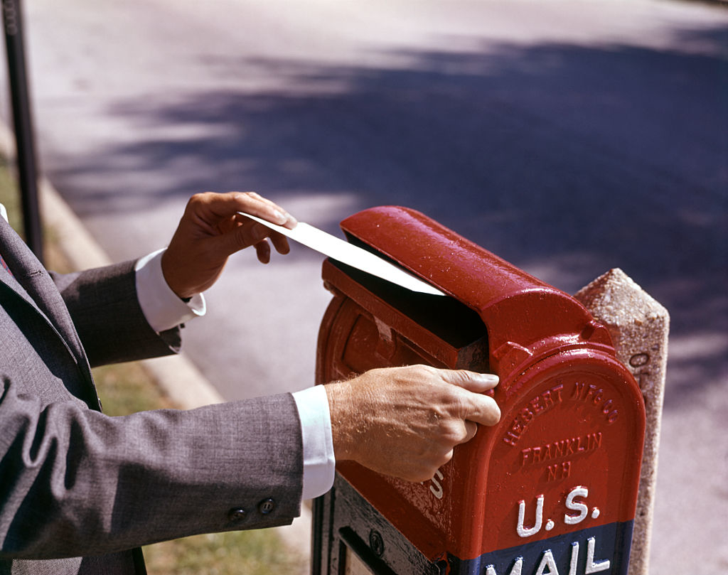 A person in a suit places a letter into a U.S. mailbox on a city sidewalk