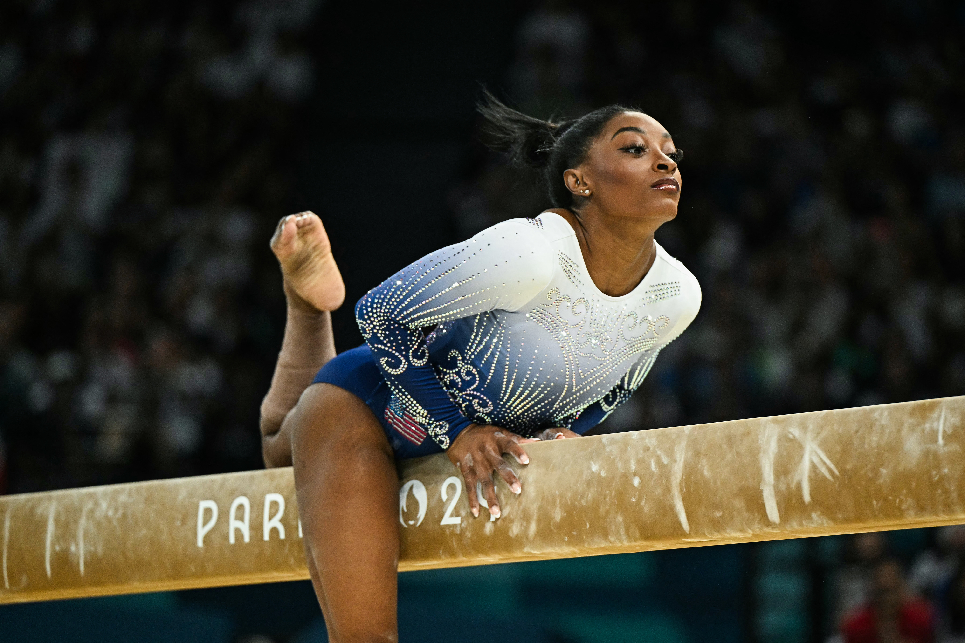 Simone Biles in a white and blue leotard performing a routine on the balance beam at the Paris 2023 event